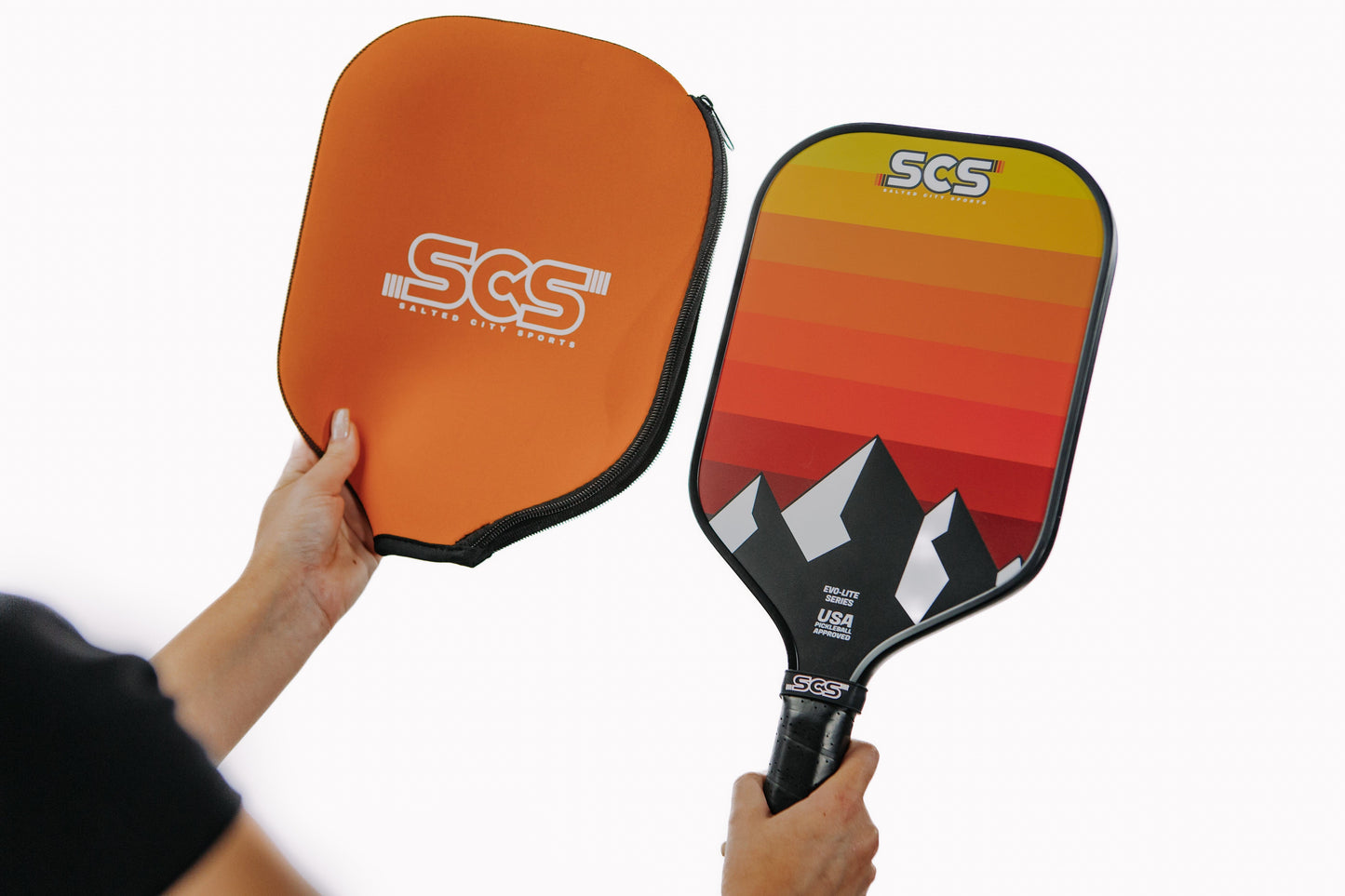 A person holds an orange Salted City Sports case in one hand and the Darkmode | Evo-Lite Series pickleball paddle, featuring a Utah Jazz-inspired mountain design with sunset stripes, in the other, both set against a white background.