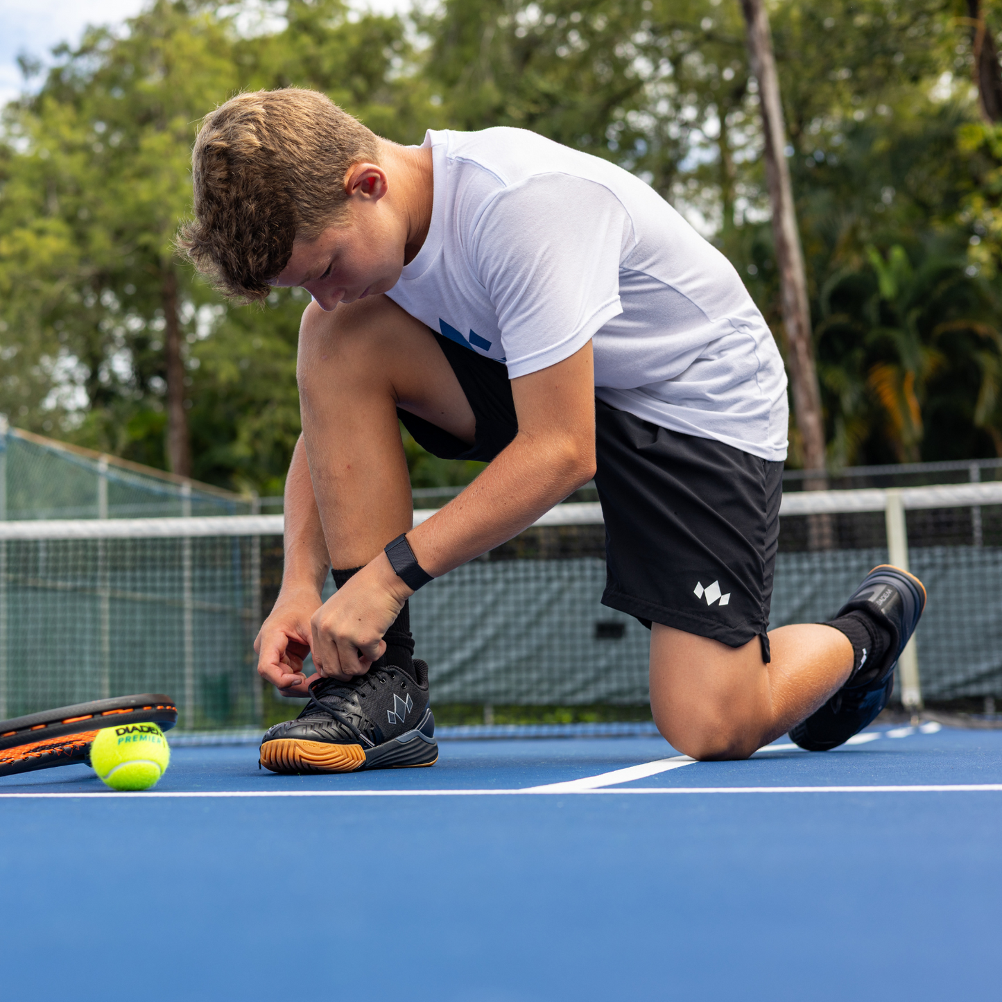 A young person kneels on a blue tennis court, tying the laces of their black Diadem Sports Diadem Court Burst shoes. Wearing a white T-shirt and black shorts, they are near two tennis rackets and a yellow ball, with green trees and a fence behind them.