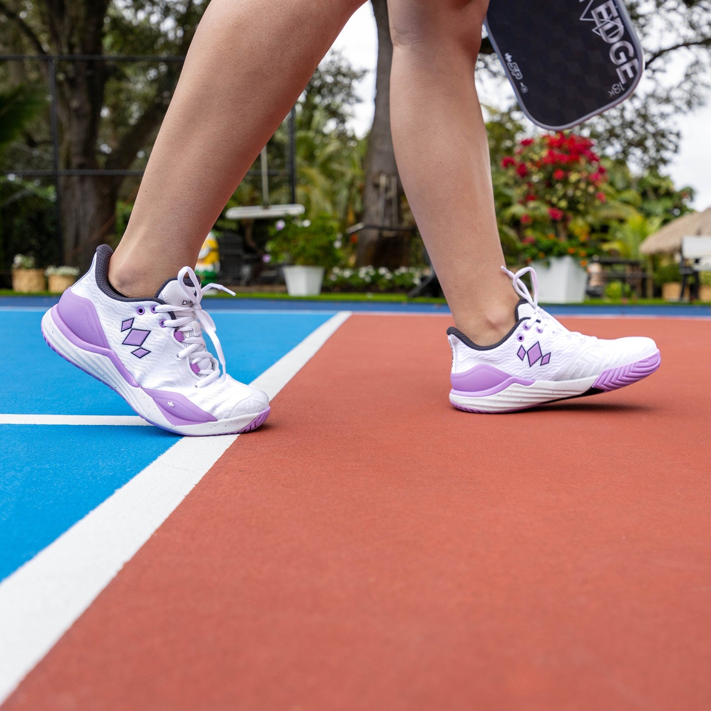 Wearing Diadem Sports' Diadem Court Burst shoes, a person steps from blue to red on a vibrant outdoor pickleball court, paddle in hand. Green trees and colorful flowers blur in the background.