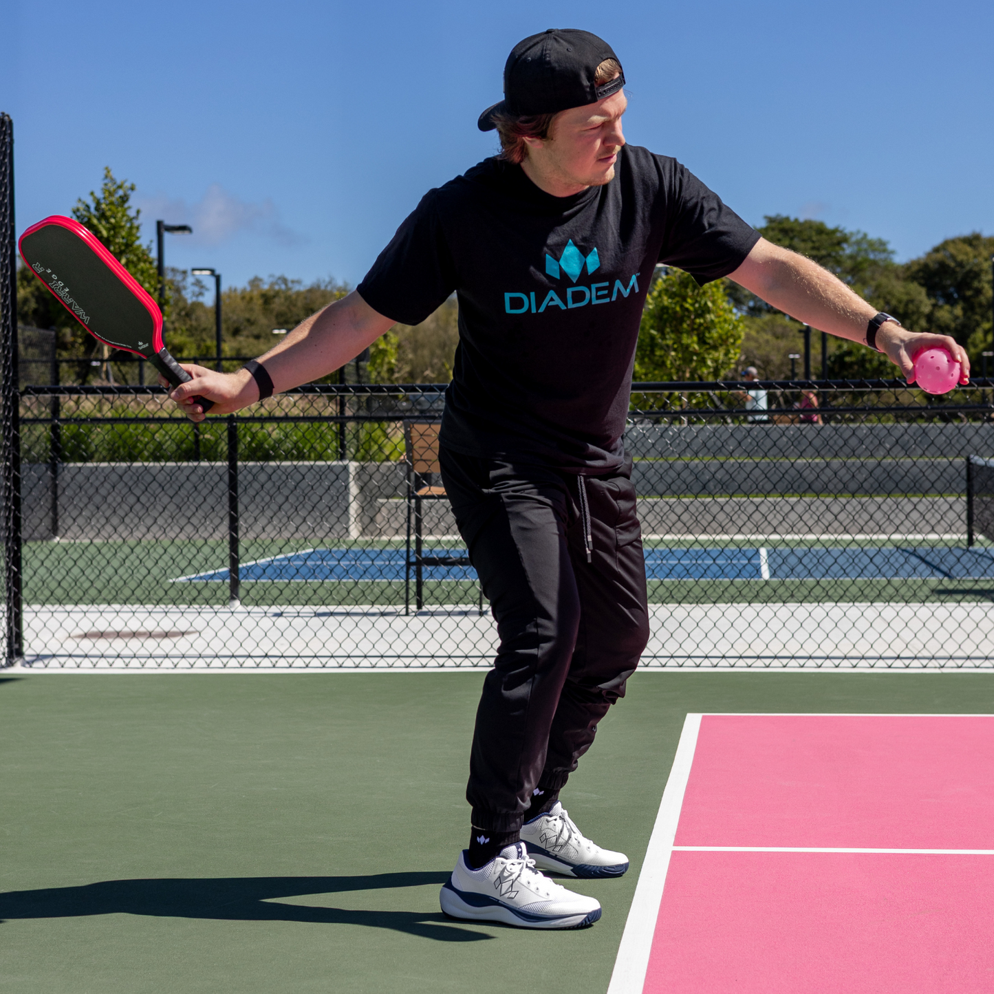 A man in a black Diadem Sports shirt and pants gets ready to serve in pickleball, holding a paddle and pink ball while wearing supportive Diadem Court Flo shoes on a court with pink and green sections, trees visible beyond the fence.