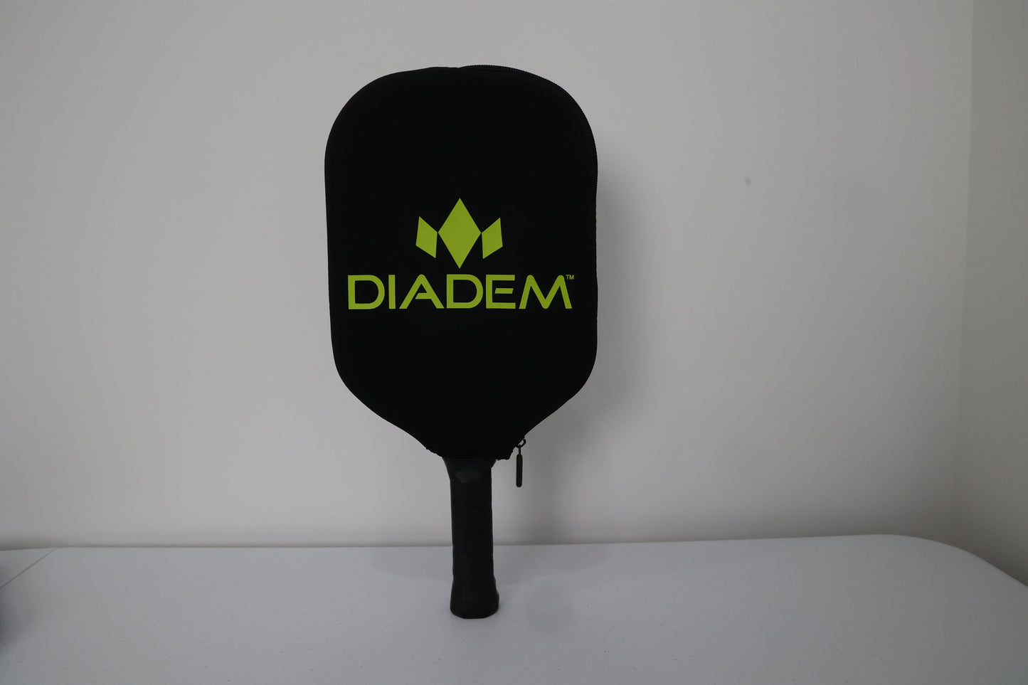 A black pickleball paddle with a Diadem Paddle Cover by Diadem Sports stands upright against a white wall. The neoprene cover features a green Diadem logo and text, while the paddle handle rests on a white surface.