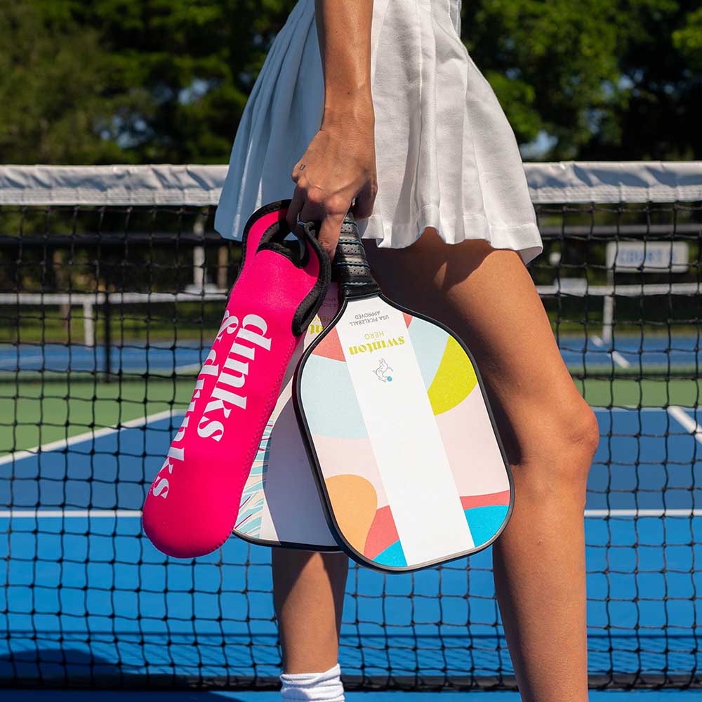 On a blue outdoor court, a person in a white tennis skirt holds Swinton Pickleball's Dinks 'N Drinks Sleeve of Pickleballs with their neoprene pickleball bag nearby, surrounded by green trees and a net in the background.