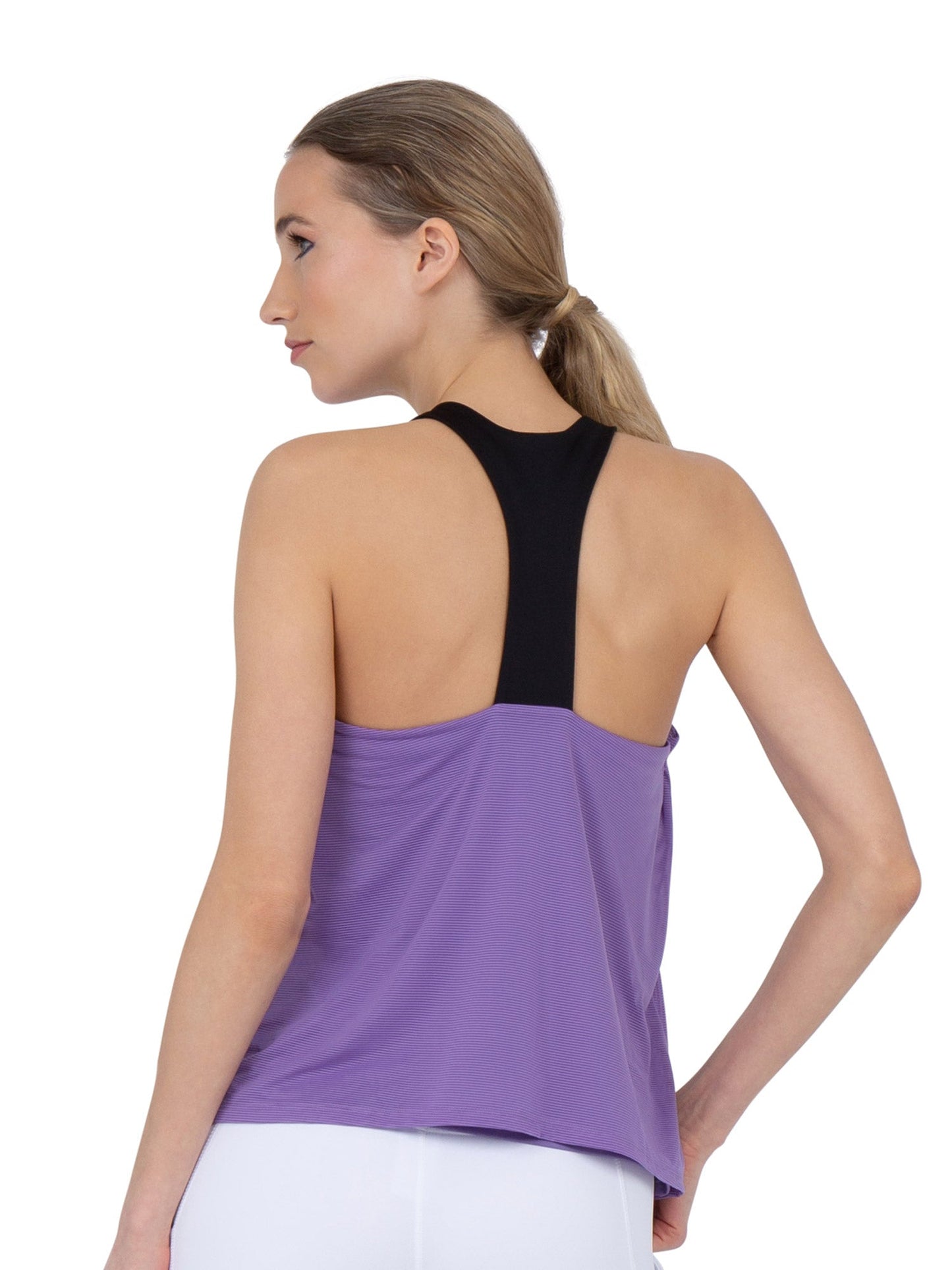 A woman with light skin and blonde hair in a low ponytail, seen from behind, wears the inPhorm NYC Double-Layered "T" Racer Back Camila Tank in Lavender/Black and fitted white pants. Her left hand rests on her hip; background is plain white.