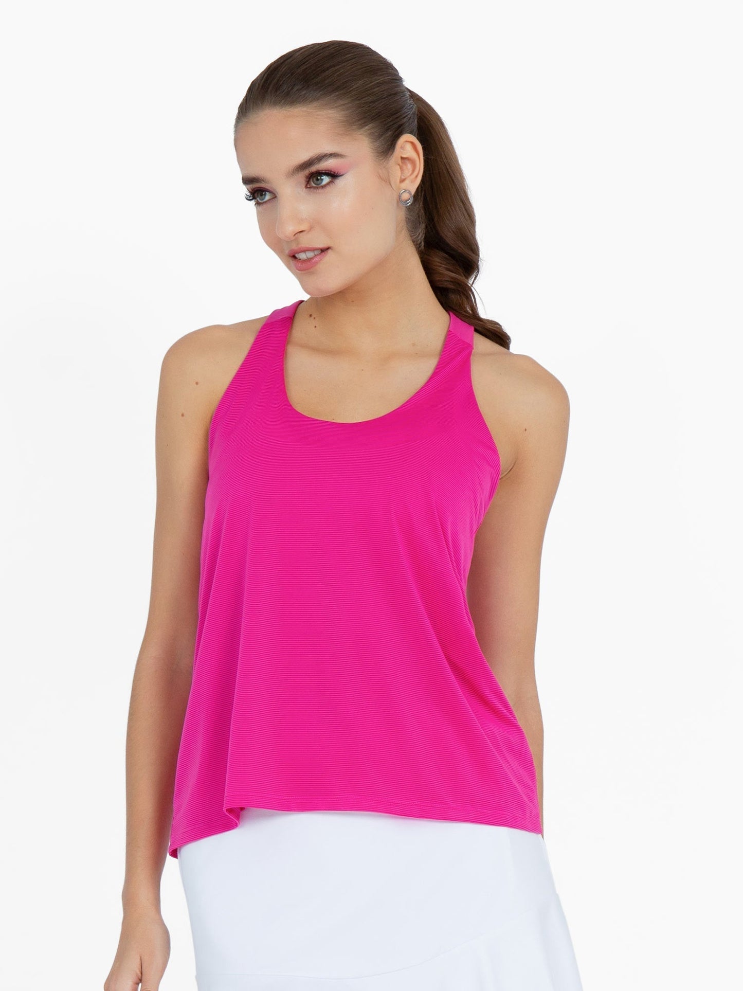 A woman with long brown hair in a ponytail wears the inPhorm NYC Double-Layered "T" Racer Back Camila Tank in Raspberry Pink and a white skirt, standing against a plain white background with a gentle expression.