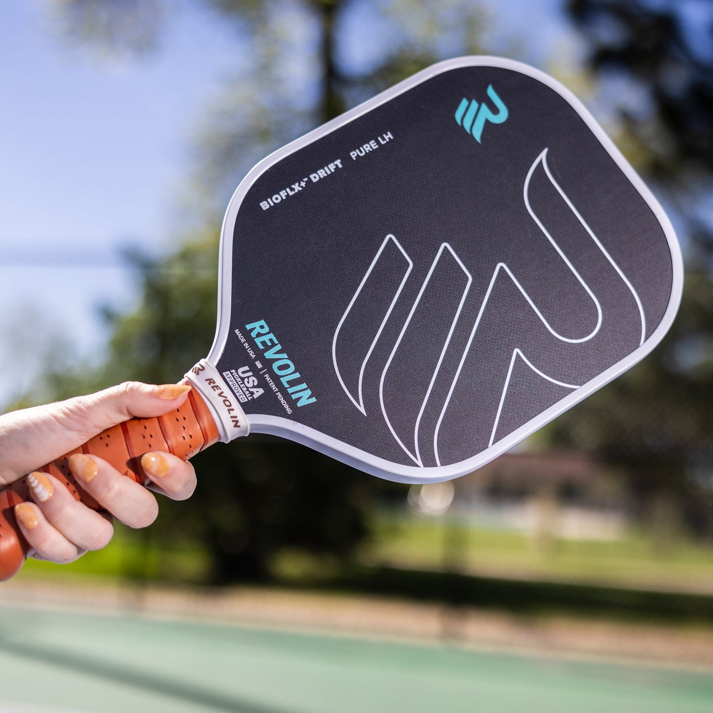 A hand with painted nails holds a black Revolin Sports Drift Pure LH Pickleball Paddle featuring a honeycomb core and white and teal accents on an outdoor court, with trees and a blurred fence in the sunny background.