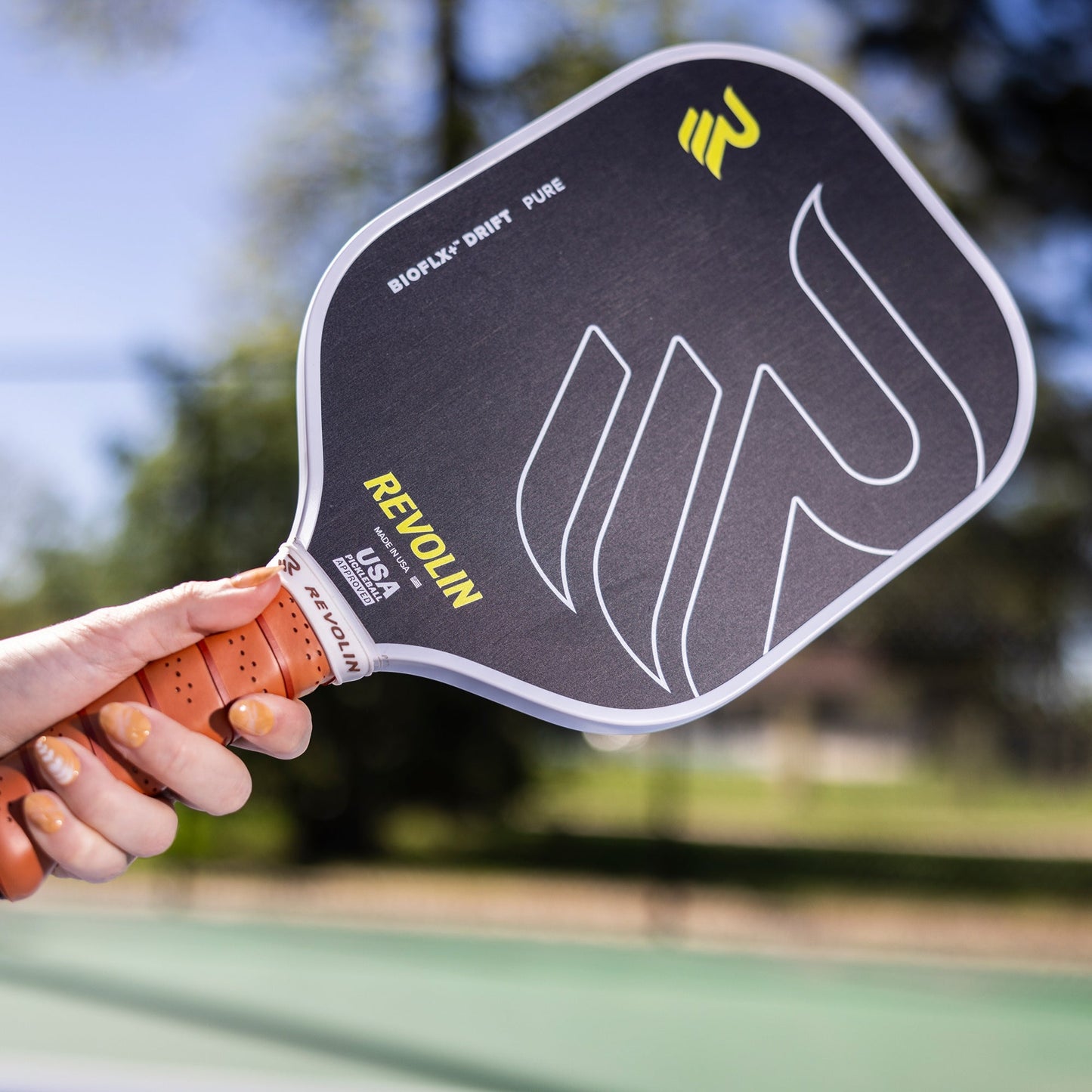 A person holds a black Revolin Sports Drift Pure Pickleball Paddle with an orange grip and bold white and yellow graphics. Designed to reduce vibration, the paddle is angled upward outdoors near blurred trees and a court.