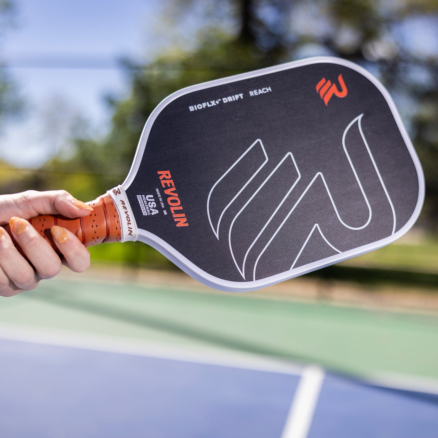 A hand with orange-painted nails holds a black Revolin Sports Drift Reach Pickleball Paddle, featuring white and orange lettering and a honeycomb core for reduced vibration, above an outdoor pickleball court with a blurred net and greenery behind.
