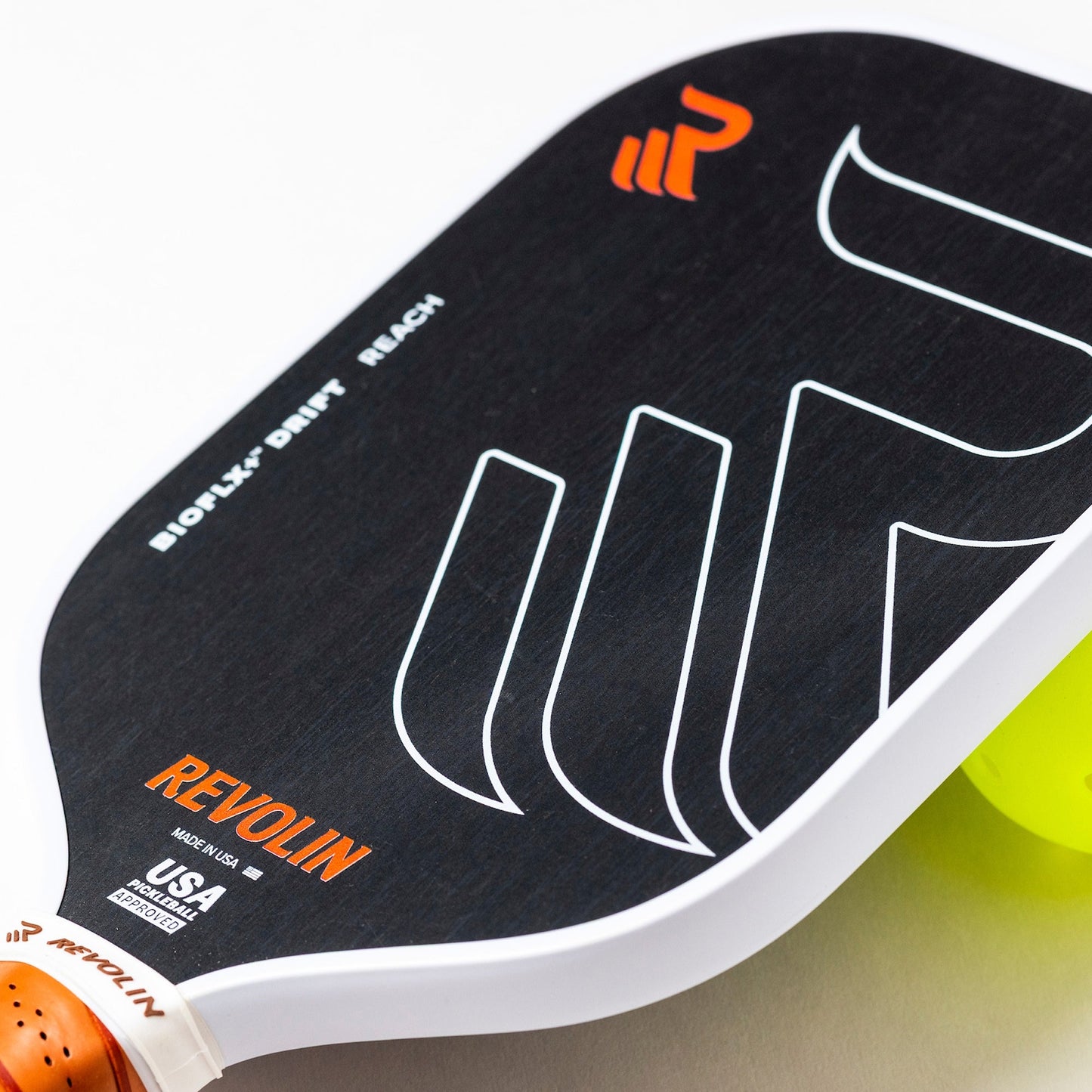 Close-up of the Drift Reach Pickleball Paddle by Revolin Sports, black with white edges, orange accents, bold graphics, and "BIOFLX+ DRIFT REACH" text; features a honeycomb core for reduced vibration. A yellow pickleball is partly visible.