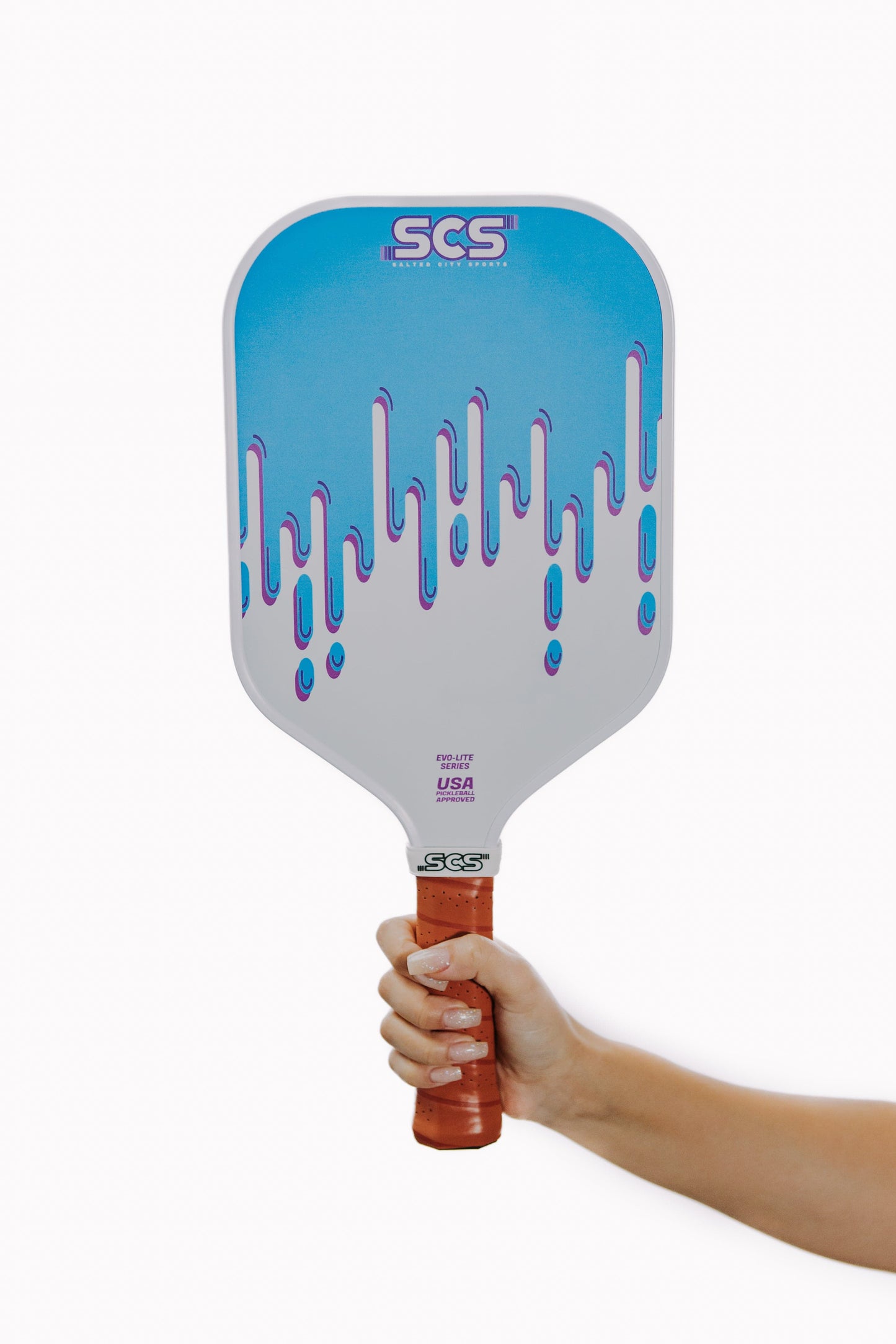 A hand holds the Salted City Sports Drippy | Evo-Lite Series pickleball paddle with a red grip, light gray face featuring blue and purple drips, SCS branding, USA Pickleball approval near the handle, and superior spin control.