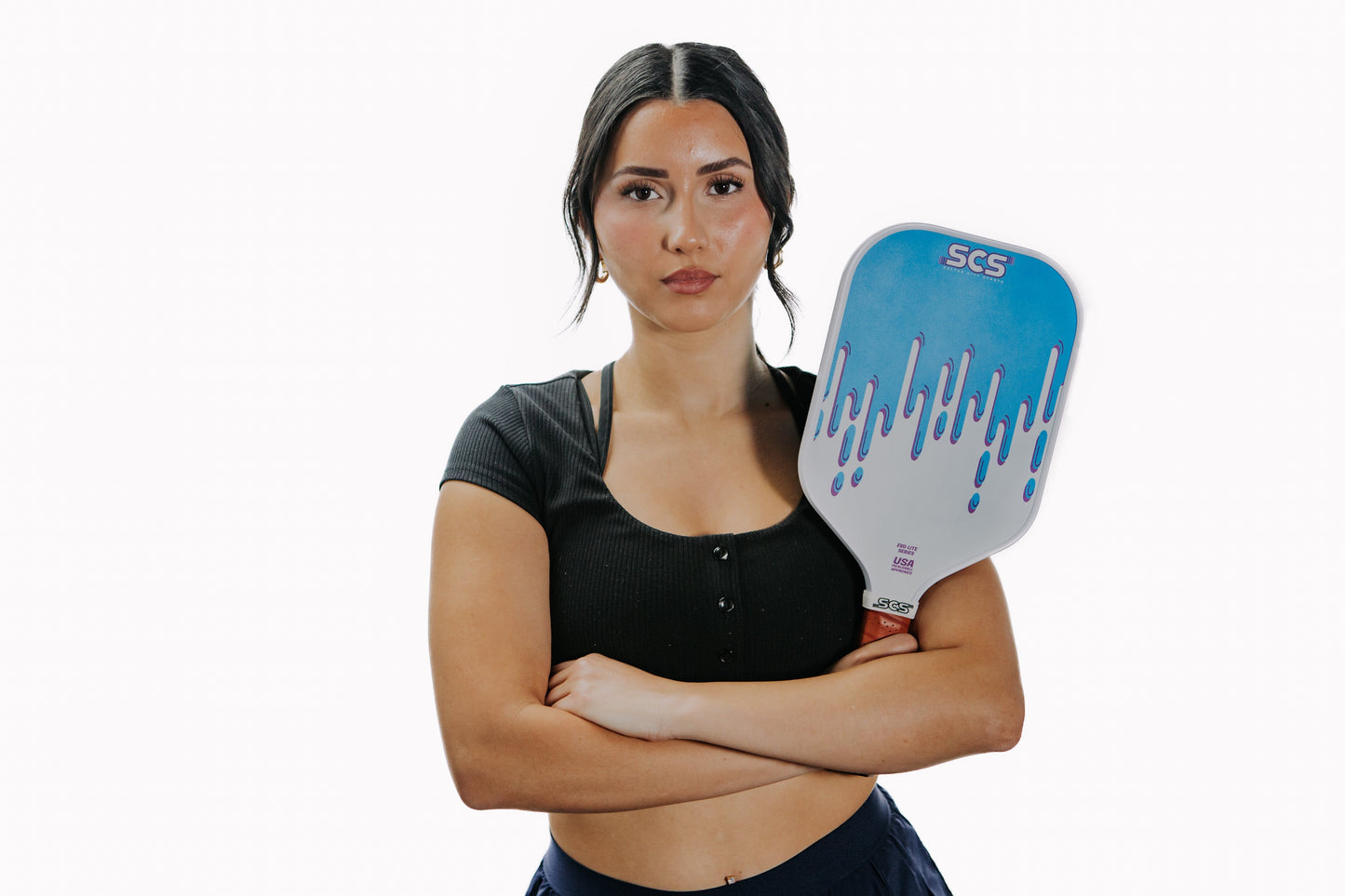 A woman with long dark hair in two low braids stands against a white background, wearing a black top and holding the Salted City Sports Drippy | Evo-Lite Series pickleball paddle, USA Pickleball approved, across her chest with a serious expression.