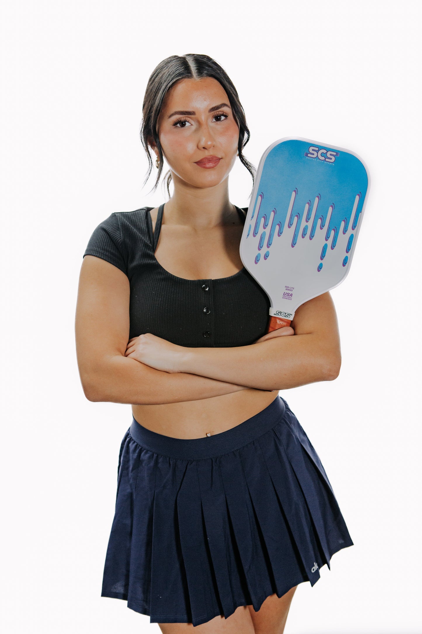 A woman with long dark hair, dressed in a black crop top and navy pleated skirt, stands confidently holding the Salted City Sports Drippy | Evo-Lite Series pickleball paddle—USA Pickleball approved—for unmatched spin control.