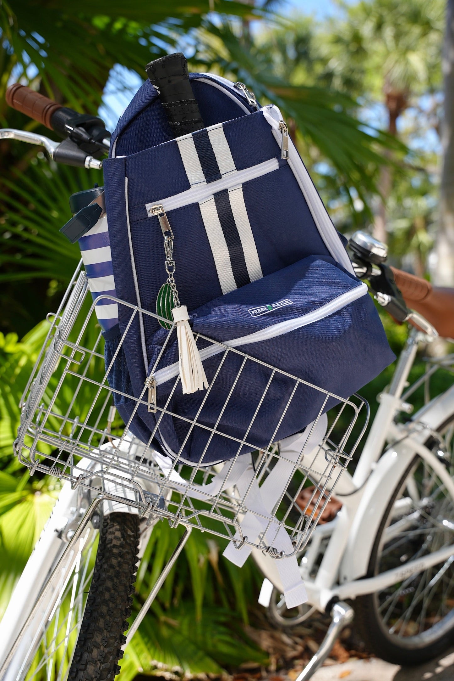 The Fresh Pickle Teddi Pickleball Backpack, navy blue with white stripes and zippers, rests in a metal bike basket on a white bicycle parked outdoors among green palm trees with sunlight filtering through the leaves.