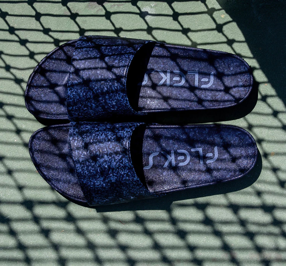 A pair of navy blue Fleks® East Beach Slide sandals by Blumaka, featuring the brand name printed upside down, are placed side by side on a green textured surface. Designed for proper alignment, ideal for athlete recovery after intense training.