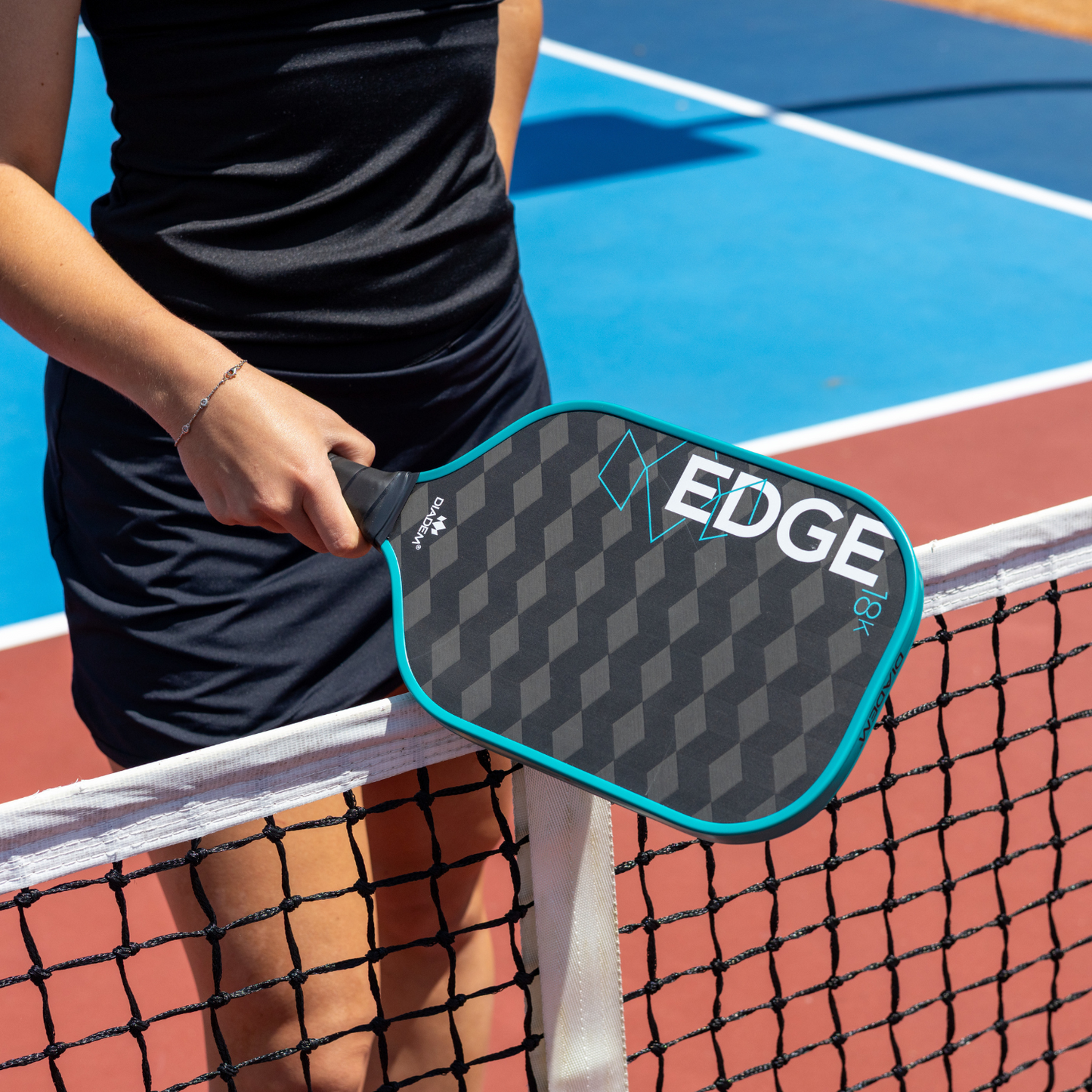 A person in a dark athletic outfit stands by a pickleball net, holding the Diadem Sports Edge 18K paddle. The black paddle features a teal border and "EDGE" text. The court surface combines blue and red colors.