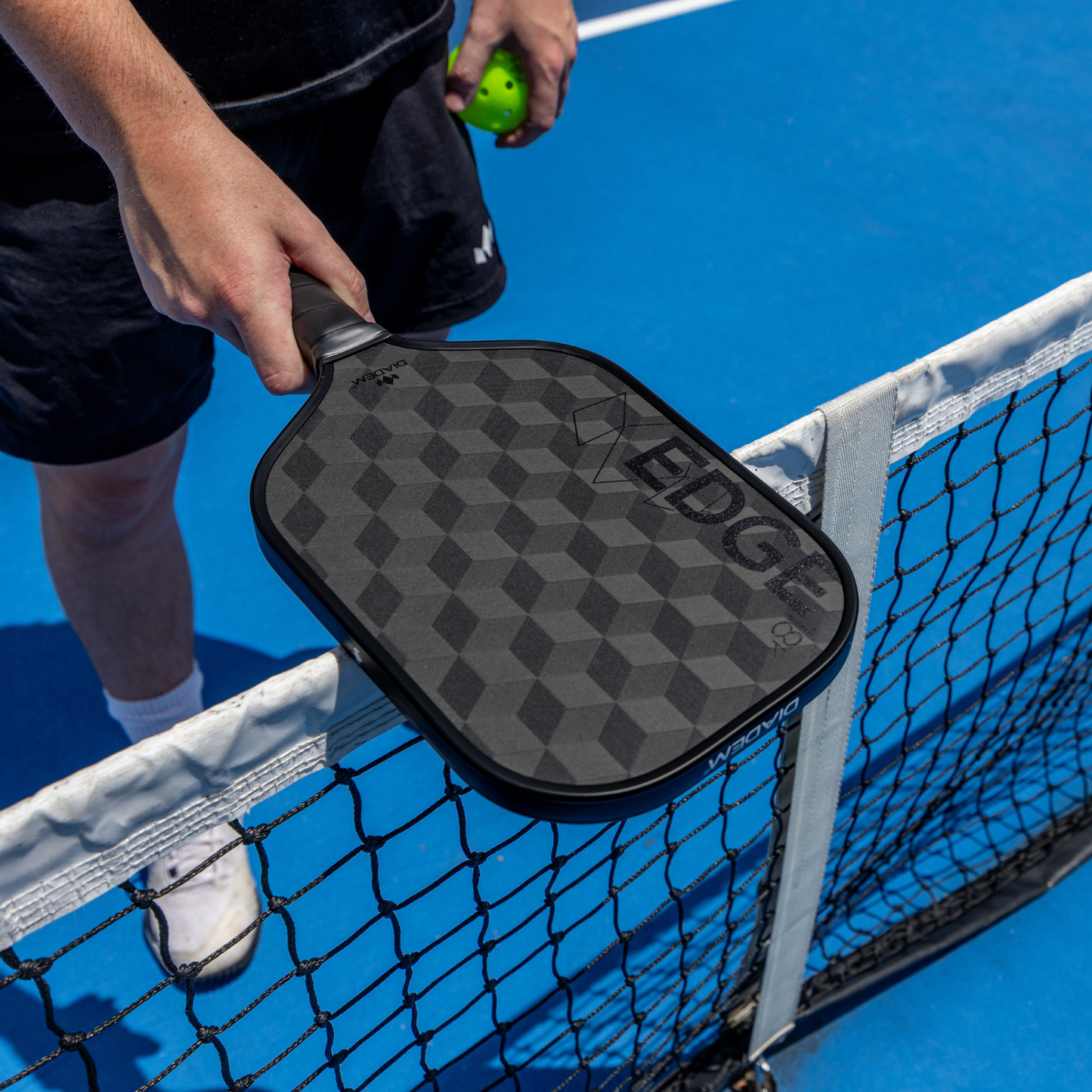 A person stands on a blue pickleball court holding a Diadem Sports Edge 18K paddle over the net. Wearing black shorts, a black shirt, and white sneakers, they hold a green pickleball in their other hand.