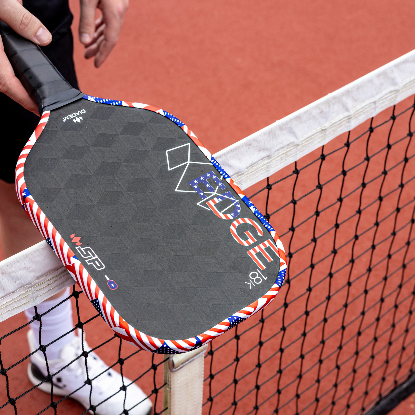 A person holds a Diadem Sports Edge 18K USAPA approved pickleball paddle with a red, white, and blue edge over an outdoor court. Only their hand, partial legs, and white shoes are visible. The paddle displays the word "EDGE.