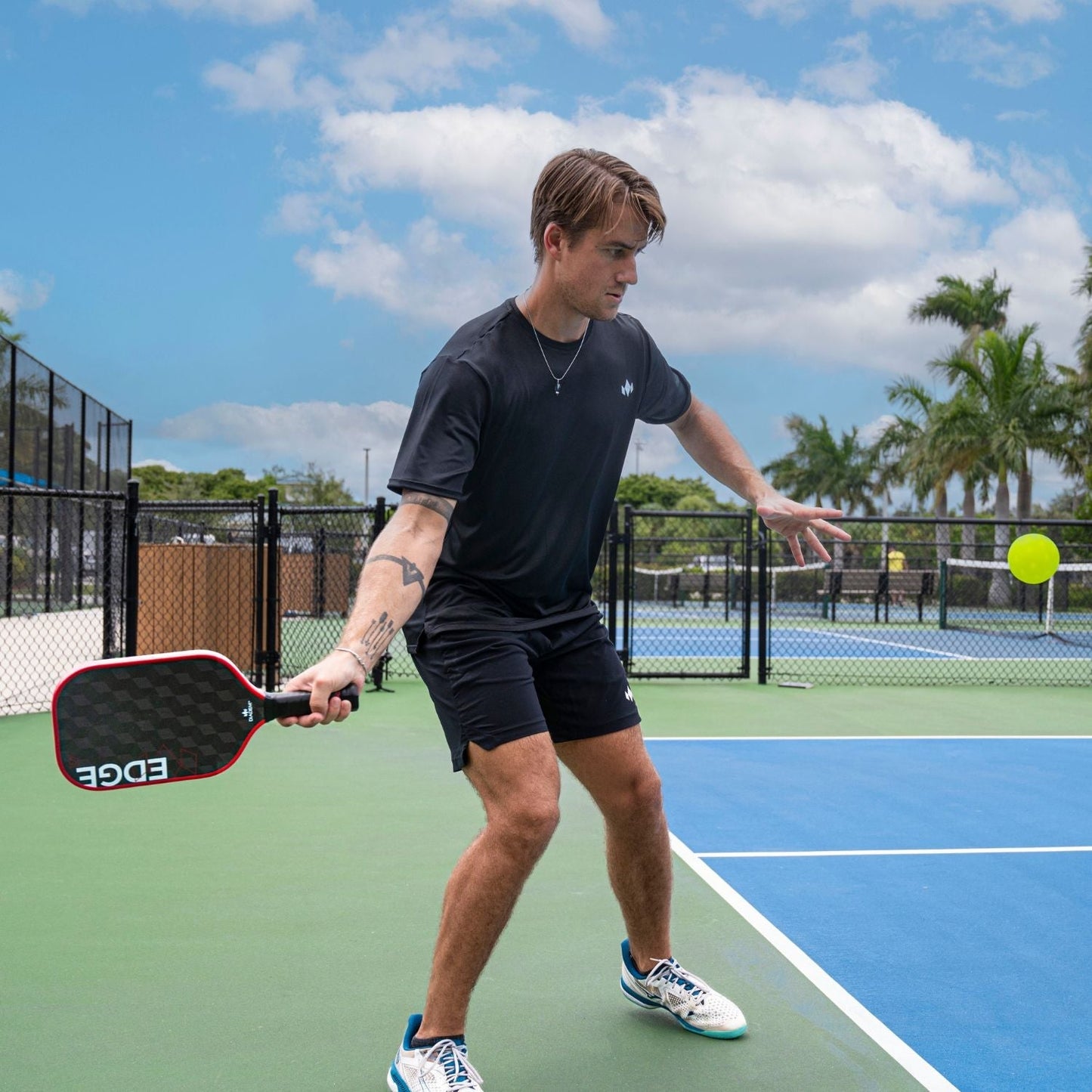 A young man in a black shirt and shorts readies to hit a pickleball with the Diadem Sports Edge 18K paddle on an outdoor court, focused as a green ball approaches beneath palm trees and a blue sky.