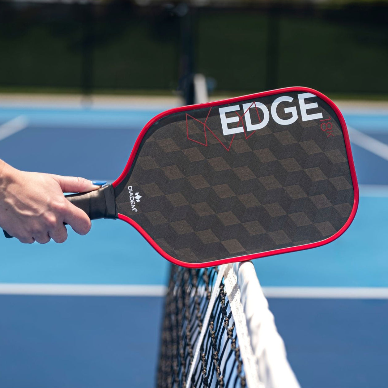 A close-up of a hand holding the Diadem Sports Edge 18K paddle, a USAPA Approved pickleball paddle, by a net on a blue outdoor court with blurred lines and fencing in the daylight.