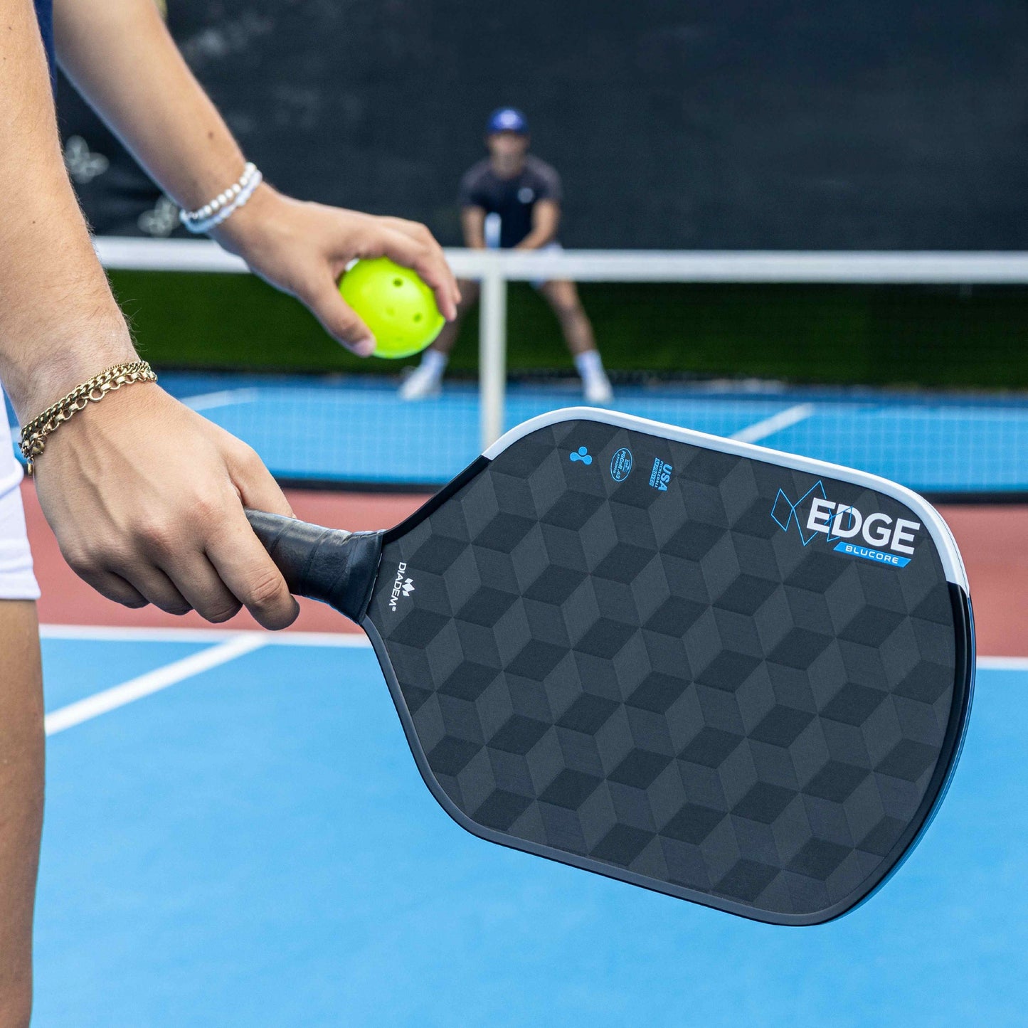 A close-up of someone holding the Diadem Sports Edge BluCore Hybrid paddle, featuring PowerSync BluCore Technology, ready to serve with a bright green pickleball on a blue and red court, while another person sits blurred in the background.