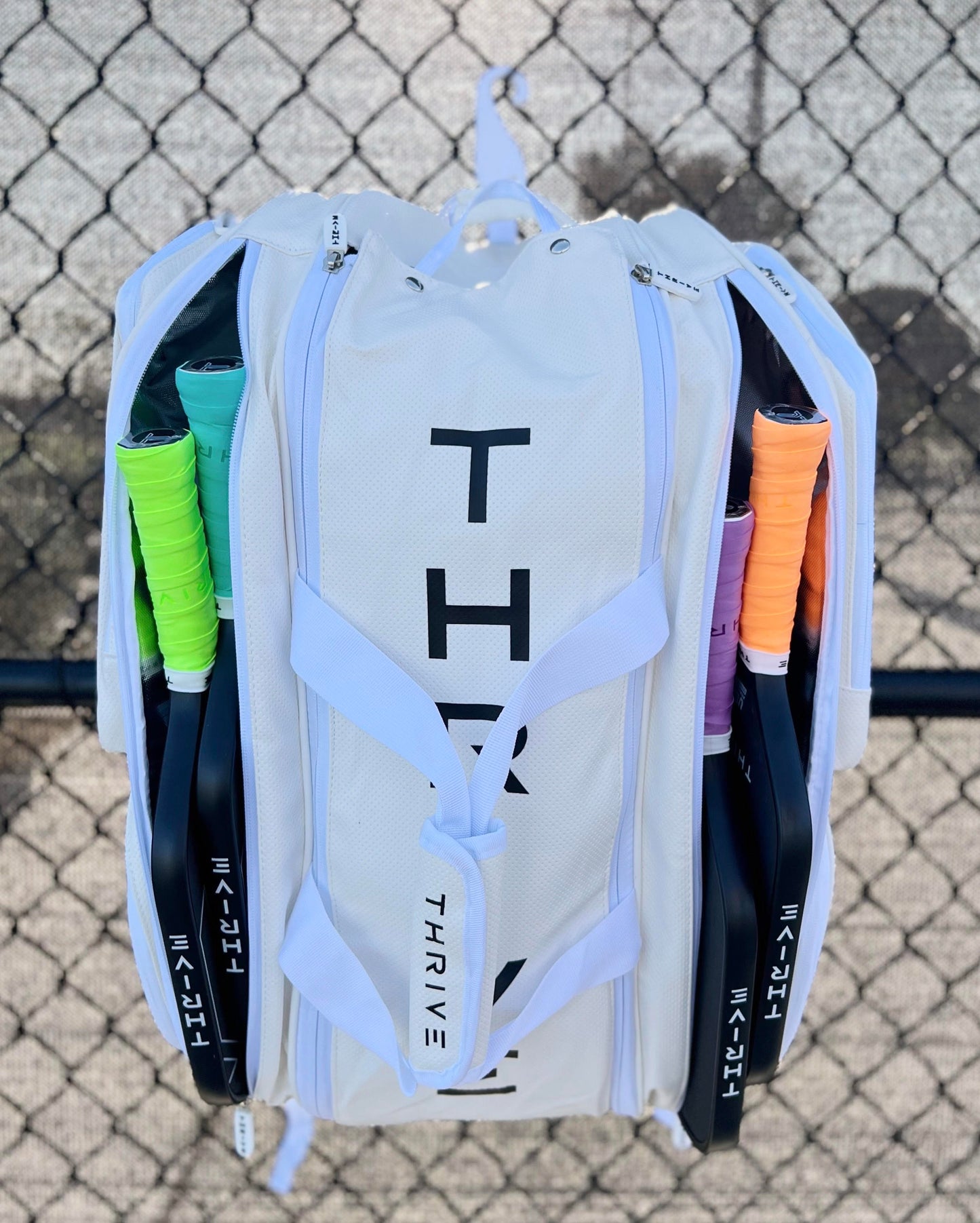 A white Elite Pro Tour Bag w/shoe bag by Thrive Pickleball hangs on a chain-link fence. Four racquets with green, purple, orange, and black grips stick out from the side pockets, handles outward. The ground is visible below.