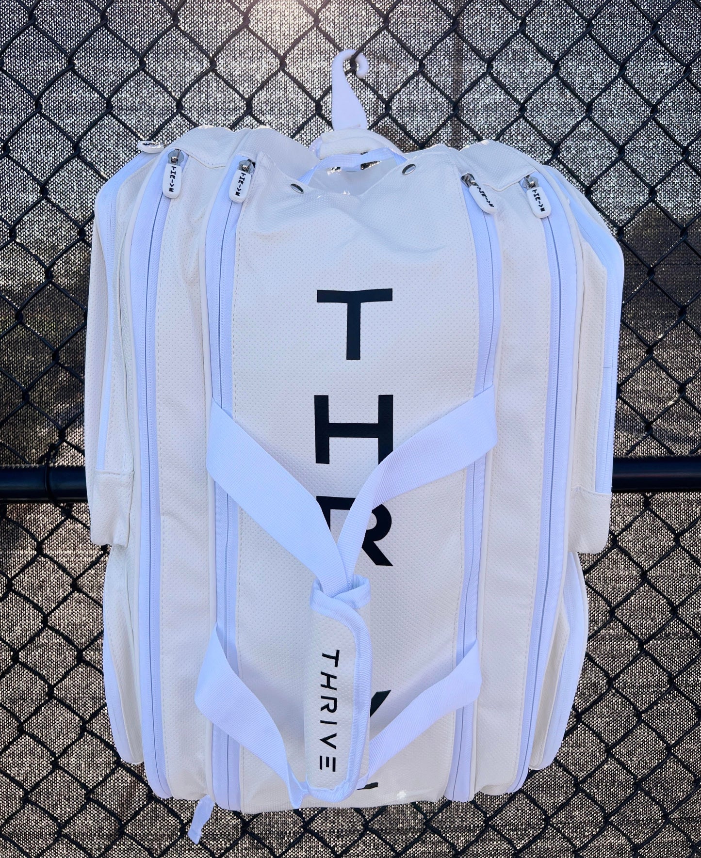 The Elite Pro Tour Bag w/shoe bag - WHITE by Thrive Pickleball hangs on a black wire fence. The large white vegan leather bag features multiple zippered compartments, white straps, and bold THRIVE lettering, with sunlight casting shadows.