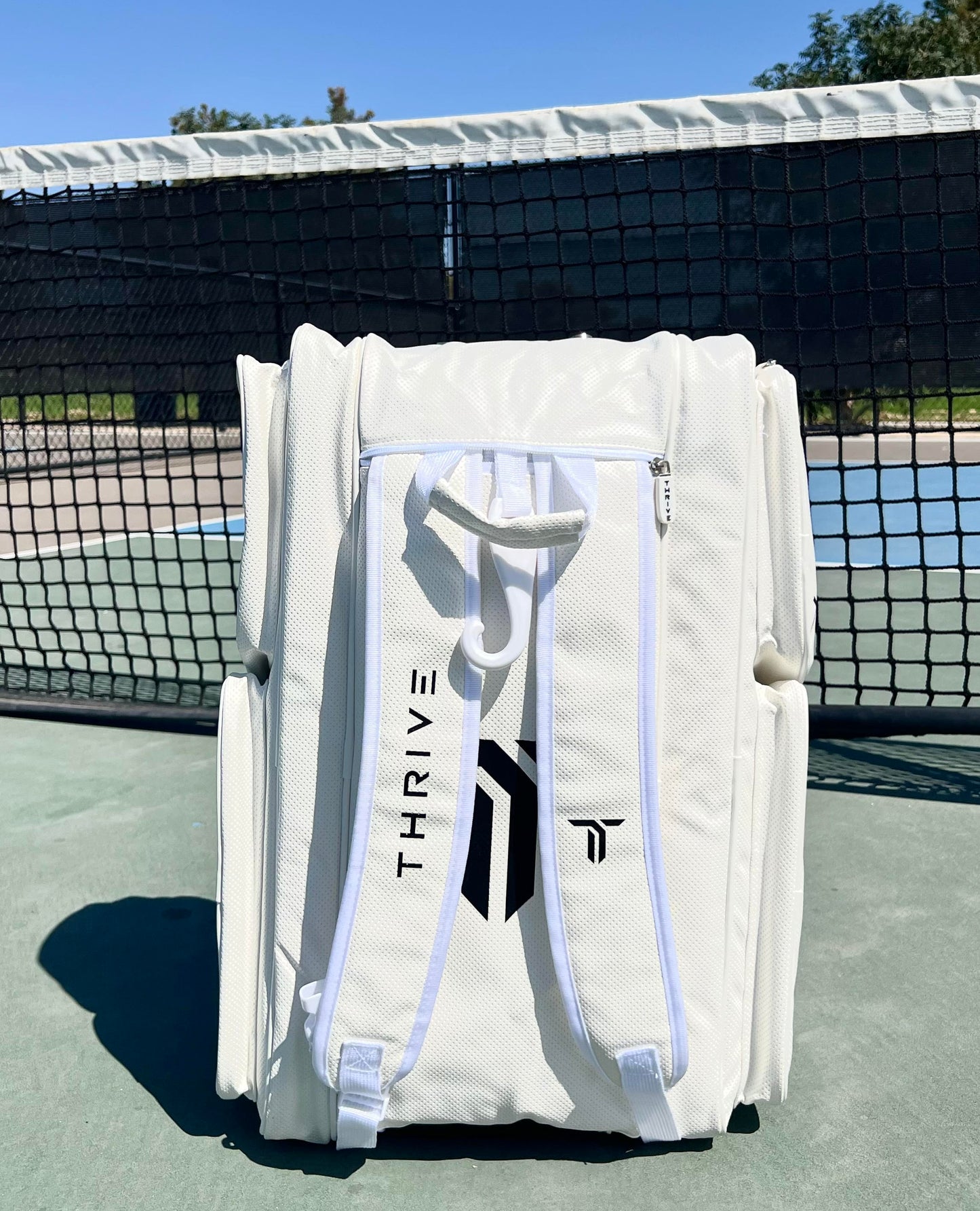 The Thrive Pickleball Elite Pro Tour Bag w/shoe bag - WHITE, made from vegan leather, stands upright on a blue tennis court near the net, featuring white zippers, handles, and “THRIVE” with a geometric logo in front.