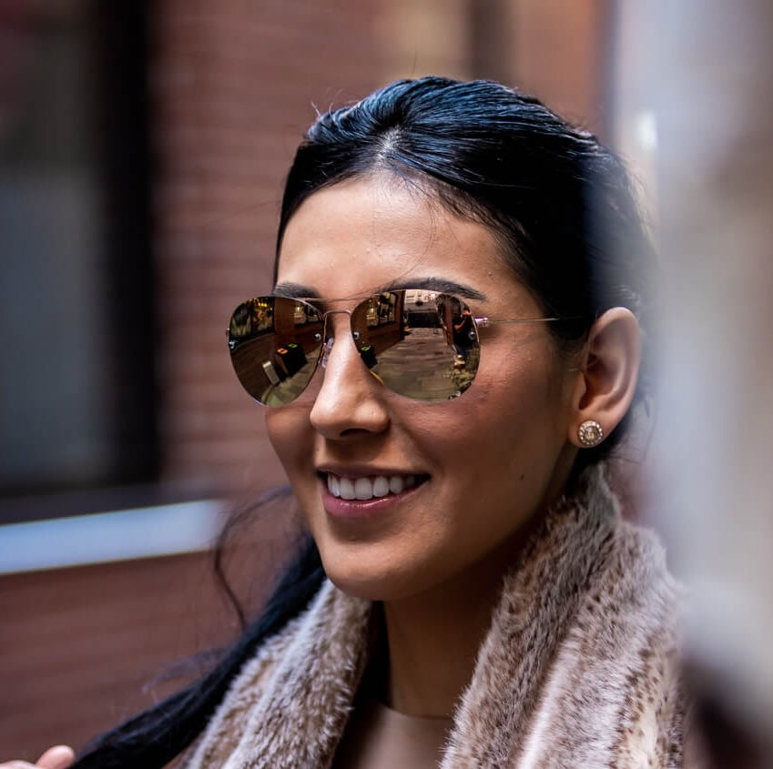 A woman with dark hair wears Epoch Eyewear’s Emerson reflective aviator sunglasses with UV protection and a light brown faux fur scarf. The mirrored lenses show buildings and a brick wall as she smiles outdoors, her face in sharp focus against a blurred background.