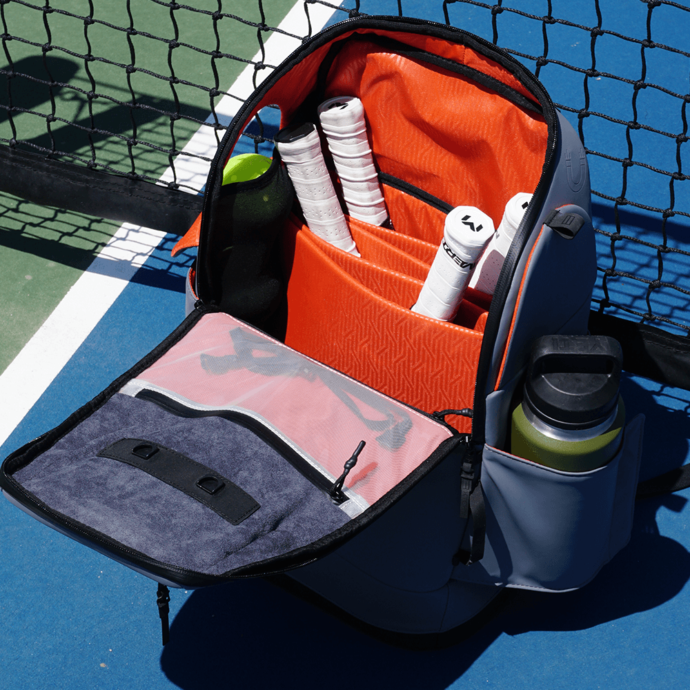 The FORWRD Court Caddy Pickleball Bag, crafted with premium materials and a vibrant orange interior, is open by a court net, displaying white racket grips, a green water bottle, and compartments for organized gear storage.