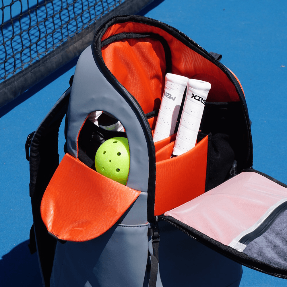 The FORWRD Court Caddy Pickleball Bag in gray and orange rests open on a blue tennis court, showing two white-handled racquets and a green perforated ball inside modular compartments. A black net appears on the left.