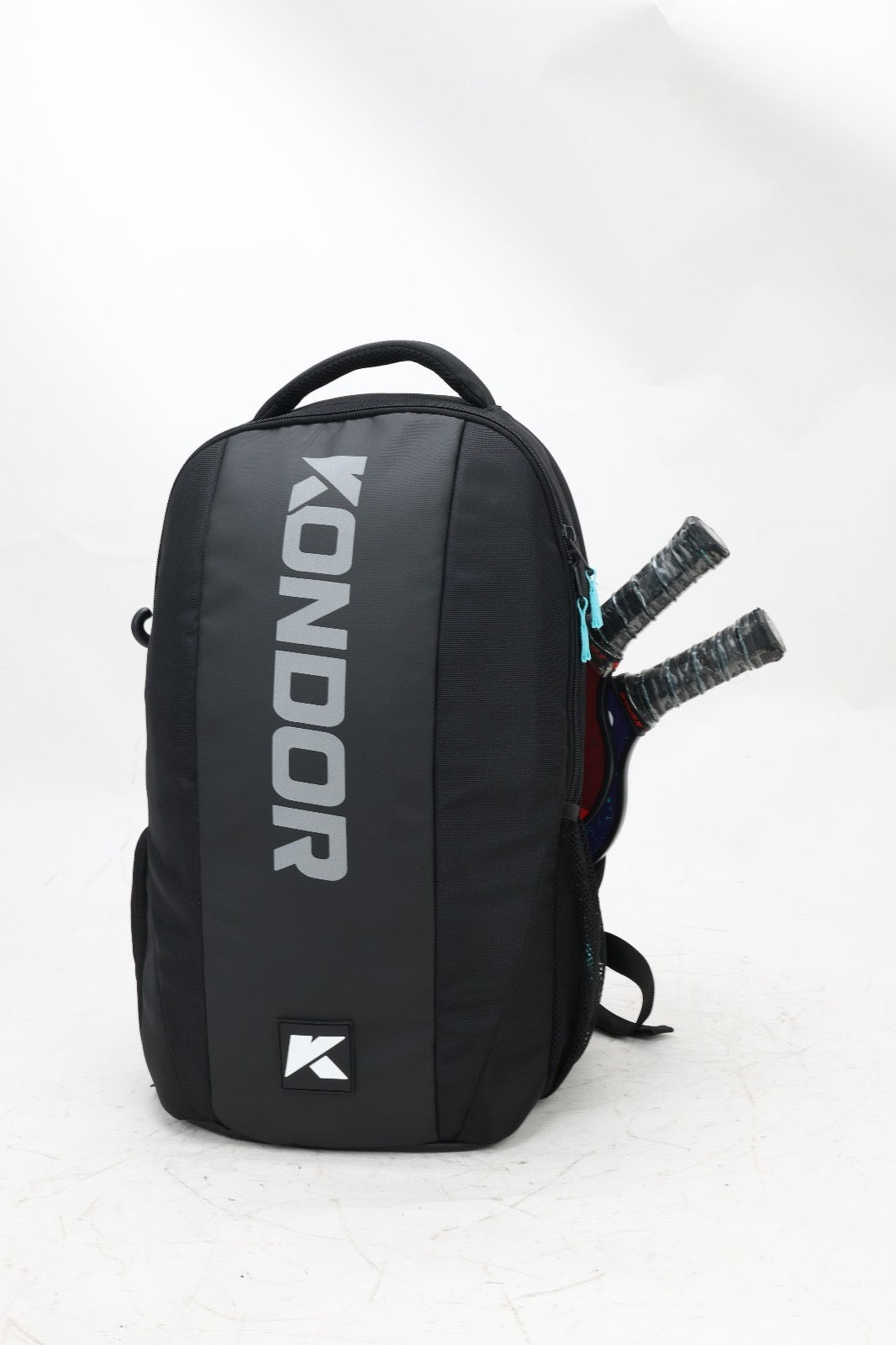 A black KONDOR PICKLEBALL Game Ready Tournament Backpack with a teal zipper and top handle stands upright. Two racket handles extend from a side pocket. The bag displays bold silver KONDOR text and a small white logo near the base.