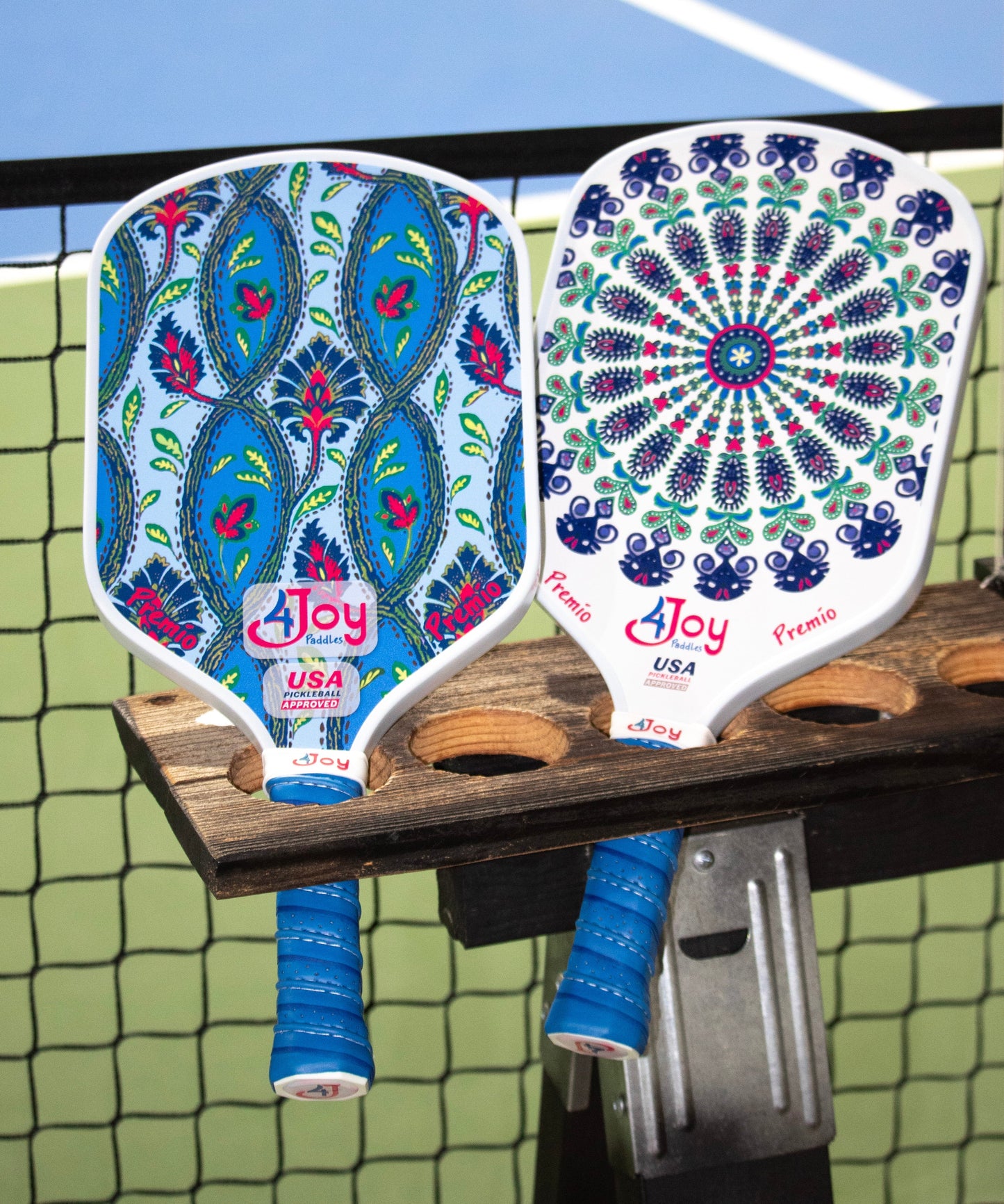 Two vibrant Fitz 4Joy Pickleball Paddle Premio Series paddles by 4Joy Paddles, with blue grip tape, rest on a wooden holder. One displays a blue and green paisley design; the other has a mandala-inspired floral pattern in blue, red, and green.