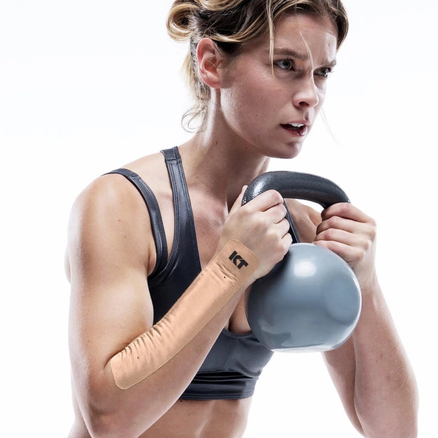 A woman with an athletic build wears KT Tape Sensitive Skin by KT Tape along her right forearm, visible while she holds a gray kettlebell to her chest. Her light hair is tied back, and the background is white.