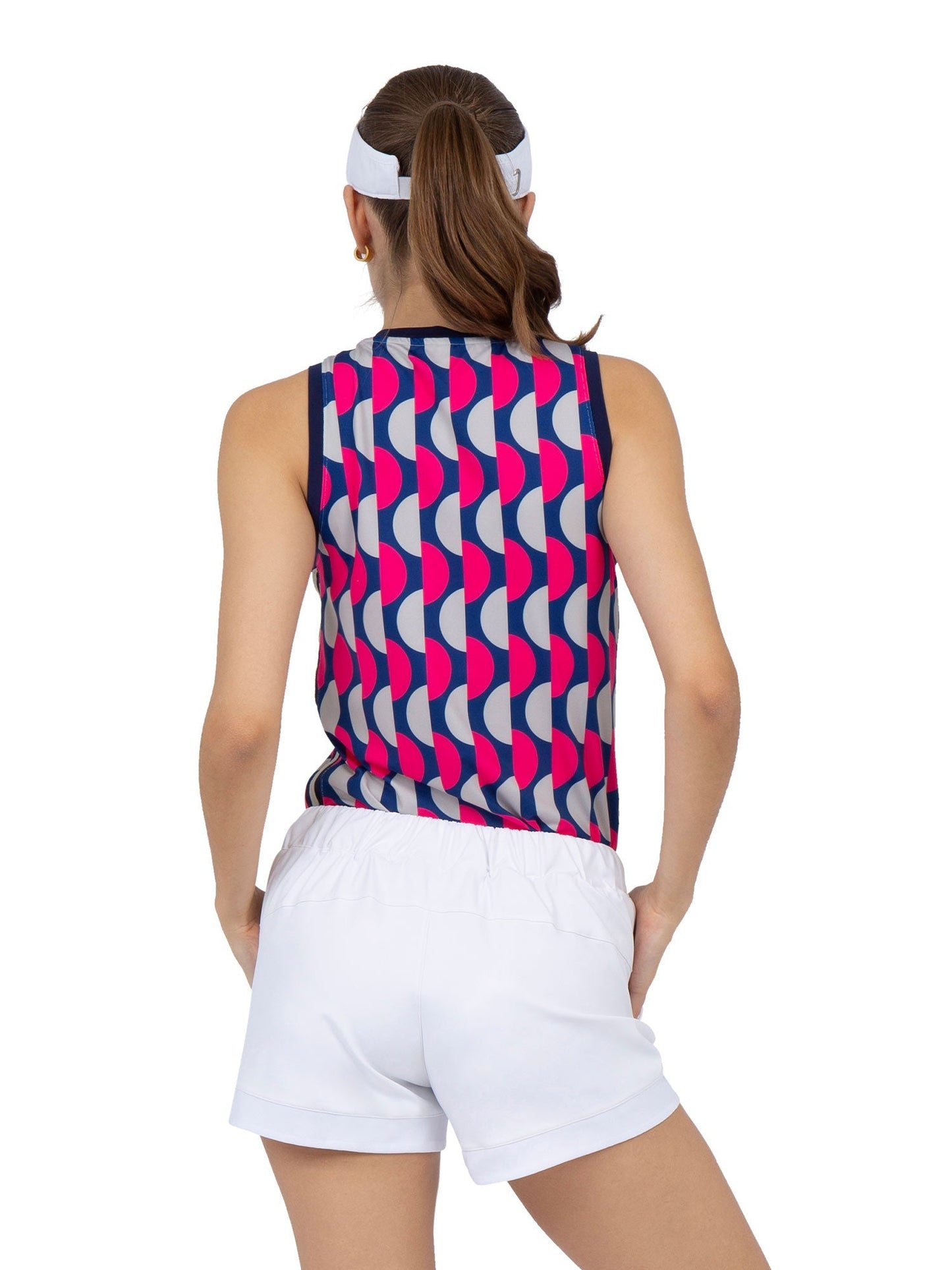 A woman with brown hair tied back stands in white shorts and a white visor, wearing the inPhorm NYC AUDREY PERFORMANCE TANK—an eco-friendly athletic top with a bold blue, red, and white geometric pattern. She faces away against a white background.