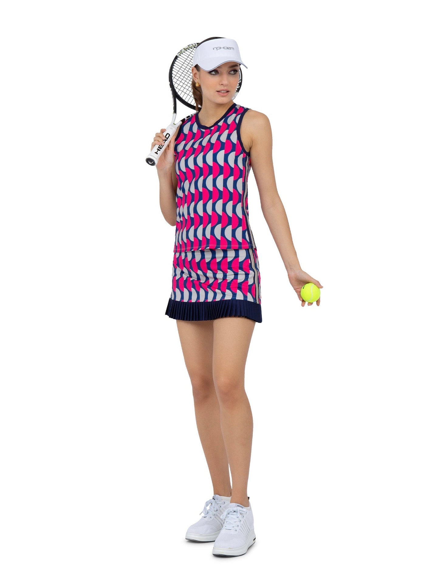 A woman wears the AUDREY PERFORMANCE TANK by inPhorm NYC, an eco-friendly, sleeveless athletic top with a geometric pink, blue, and white pattern. She holds a tennis racket and ball; her UPF 50+ outfit stands out against a white background.