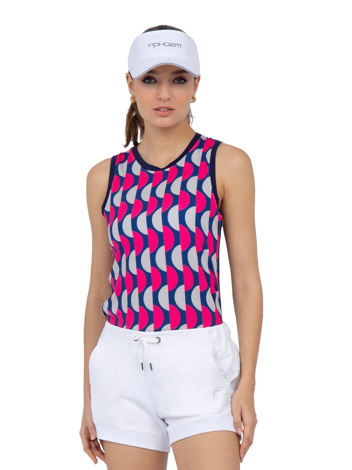 A woman models the inPhorm NYC AUDREY PERFORMANCE TANK, an eco-friendly athletic top with a bold blue, red, and white geometric pattern and UPF 50+ protection. She pairs it with a white visor and drawstring shorts against a plain background.