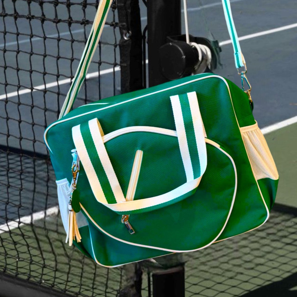 The Fresh Pickle Lola Pickleball Tote Bag in green with white trim and yellow accents hangs on a net post. With dual zippers, front pocket, striped handles, and shoulder strap, it’s perfect for carrying pickleball gear. Green and blue court in background.