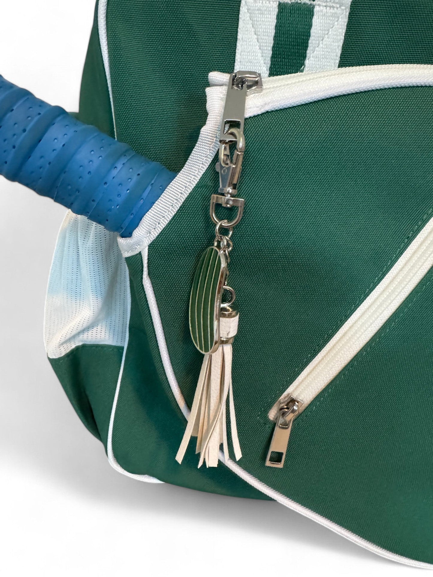 The Fresh Pickle Lola Pickleball Tote Bag is green with white zippers and trim, shown with a blue tennis racket handle, plus a zipper pull keychain featuring a green-striped racket charm and white leather tassel.