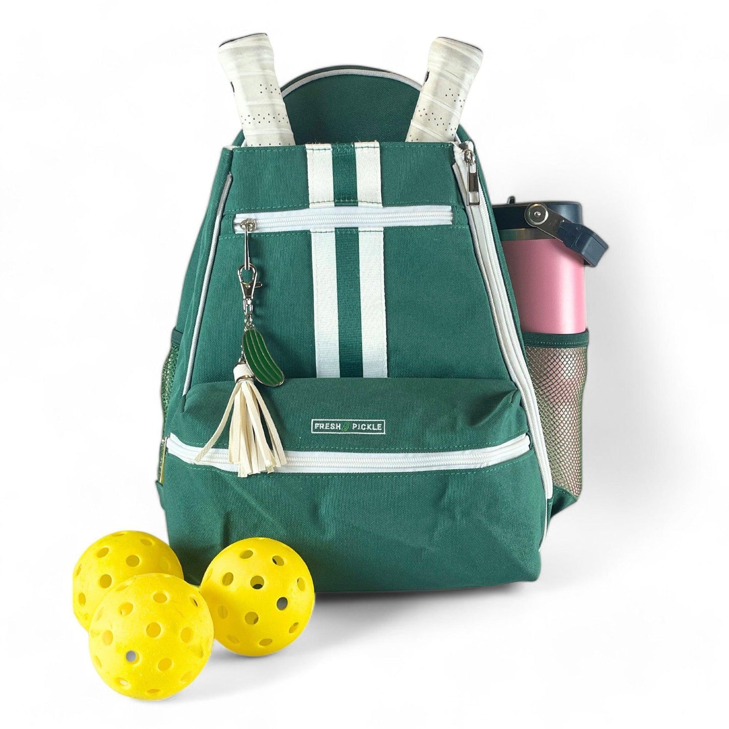 The Fresh Pickle Teddi Pickleball Backpack, featuring green and white stripes, holds two paddles, a keychain, and a white tassel. A pink water bottle fills the side pocket while three yellow pickleballs rest in front.