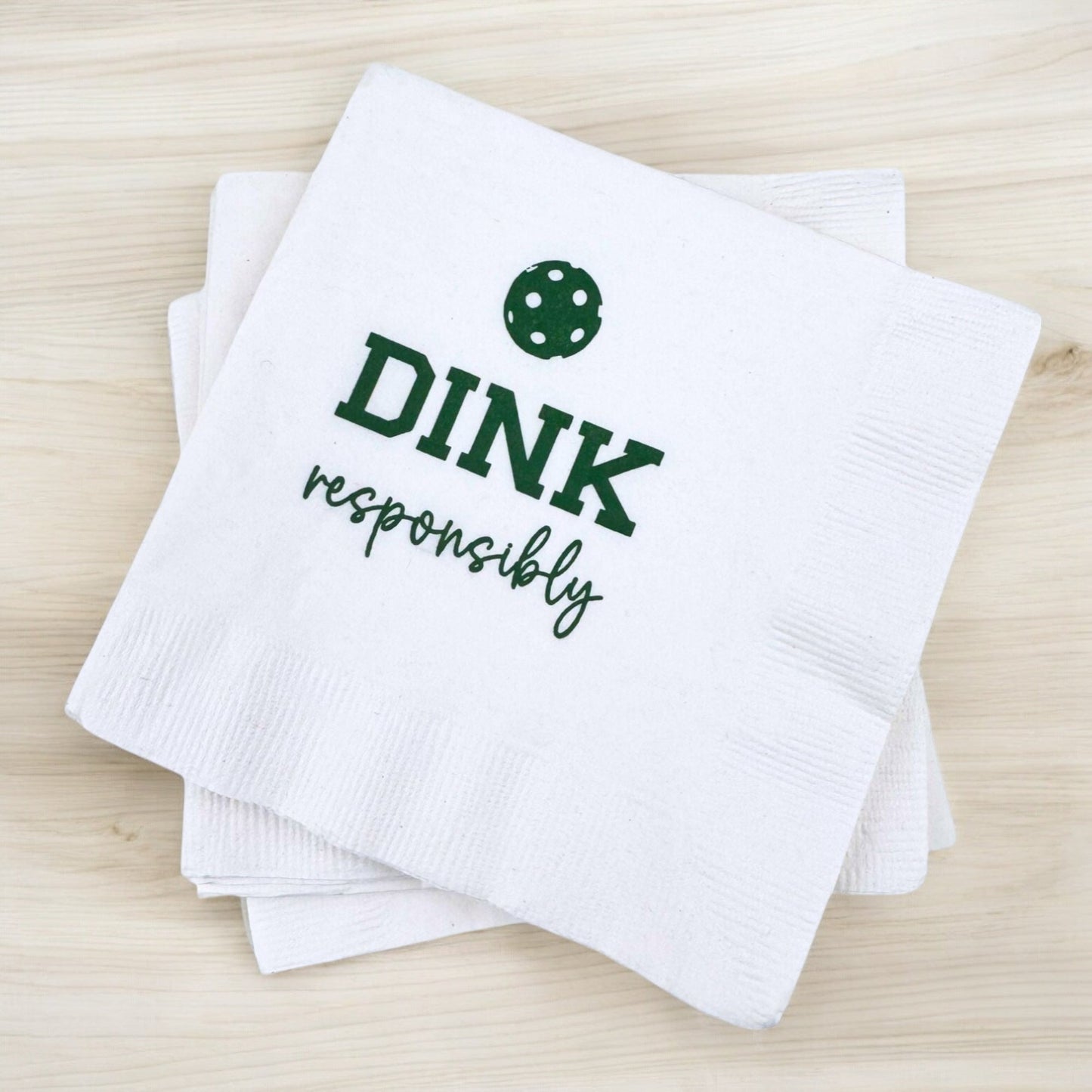 Fresh Pickle's Pickleball Cocktail Napkins rest on a light wood surface, showing a green pickleball and "DINK responsibly" in bold and script. Two more themed napkins from the set are partly visible underneath.