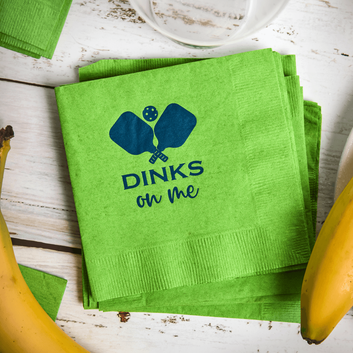 Fresh Pickle Pickleball Cocktail Napkins in bright green sit on a rustic white table. They feature blue paddle and ball graphics above "DINKS on me," with bananas and part of a white bowl nearby.
