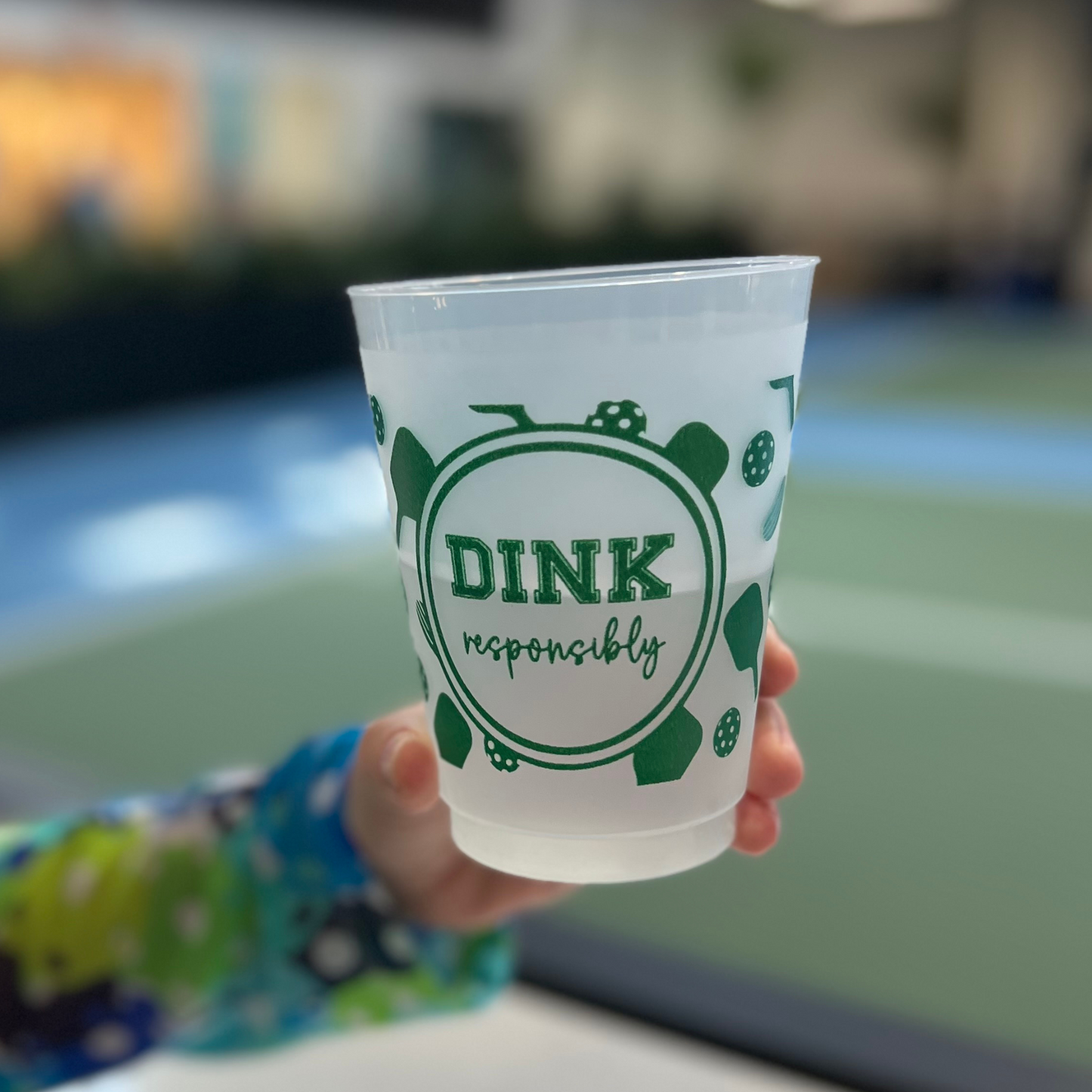A hand holds a shatterproof Pickleball Roadie Cup by Fresh Pickle, featuring green "DINK responsibly" text with pickles and a paddle. In the blurred background, an indoor pickleball court is visible.