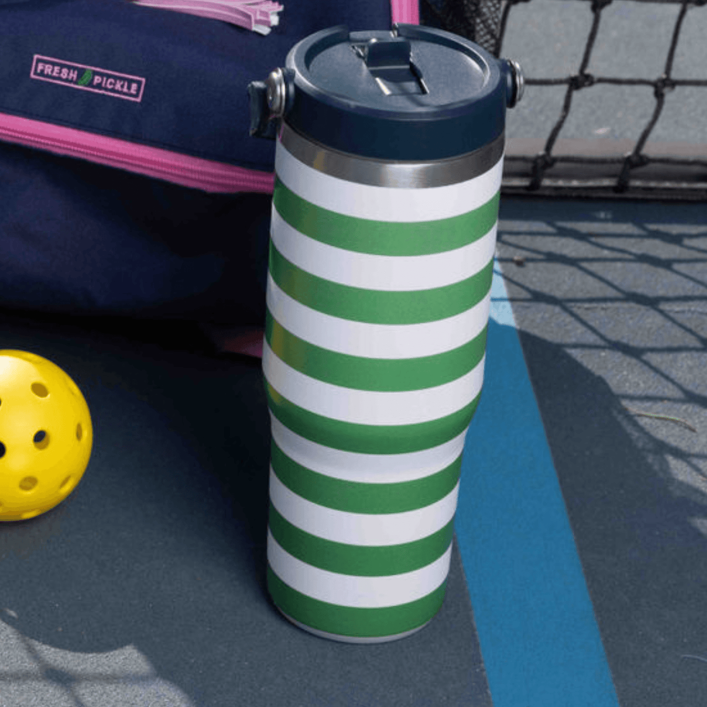 The Fresh Pickle Tumbler by Fresh Pickle, a green and white striped 30 oz double-walled vacuum bottle with a navy lid, rests on a tennis court by a yellow pickleball, a navy bag with pink zipper, and metal fence shadows.