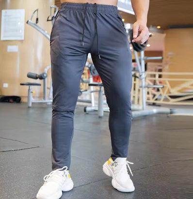 A person wears DYM Athletics Men's Luxury Lounge Jogger in dark gray, featuring zipper pockets, with white athletic shoes in a gym. Gym equipment and wooden walls are in the background; black rubber matting covers the floor. Face not visible.