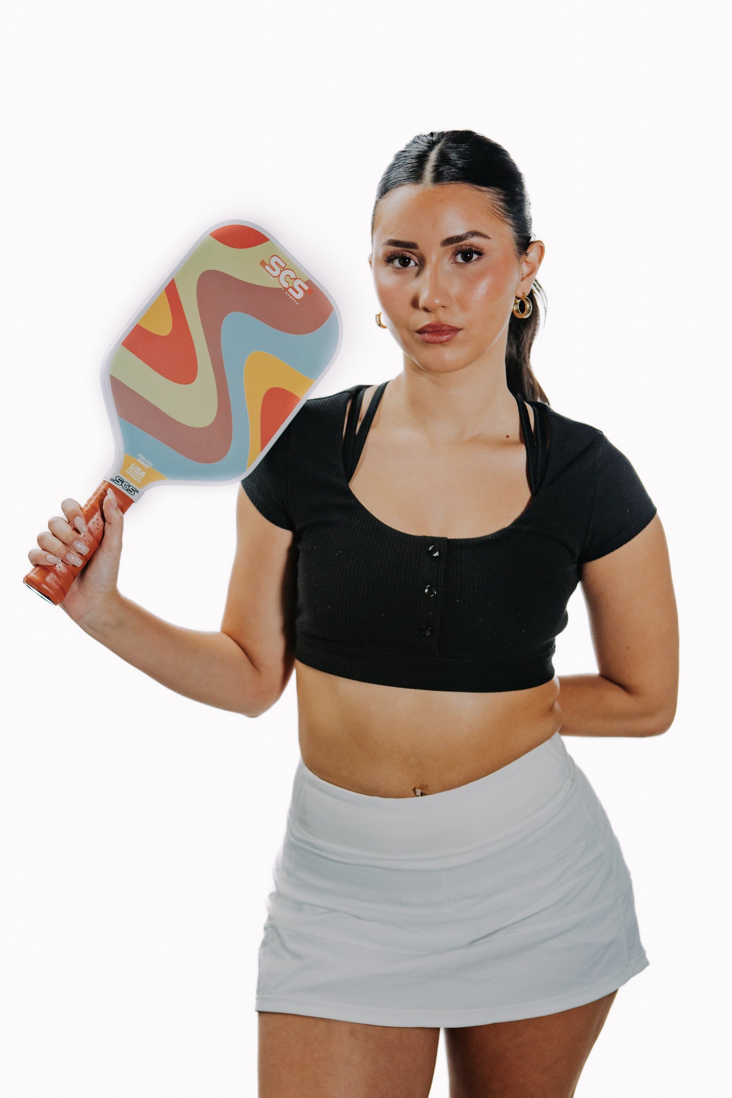 A young woman with long dark hair in a ponytail holds the Salted City Sports Groovy | Evo-Lite Series pickleball paddle featuring a retro swirl design. She wears a black crop top and white athletic skirt, standing confidently against a plain white background.