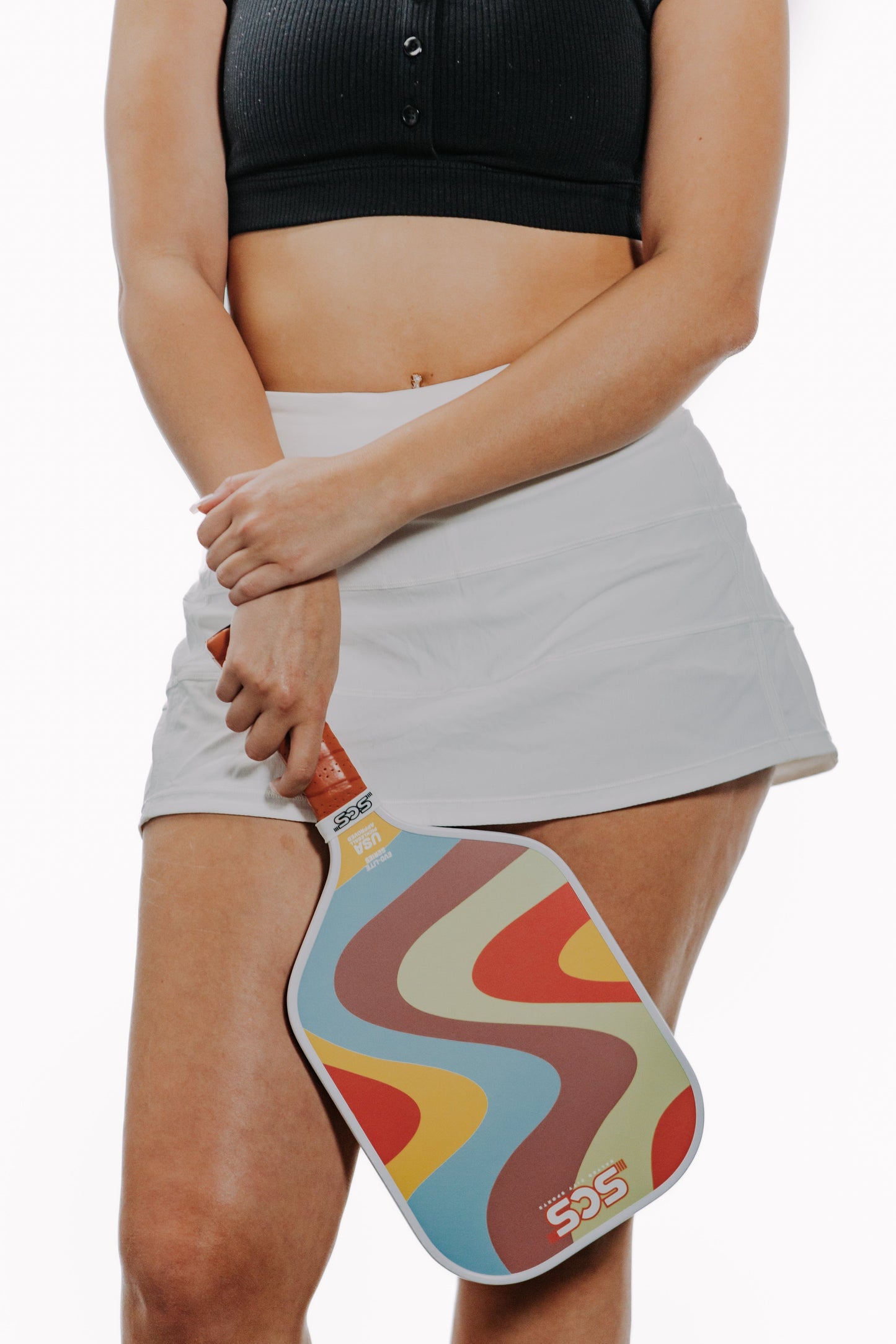 A woman in a black crop top and white skirt holds the Salted City Sports Groovy | Evo-Lite Series paddle, featuring red, yellow, and blue wavy patterns. She crosses her arms as the USA Pickleball-approved paddle rests by her leg.
