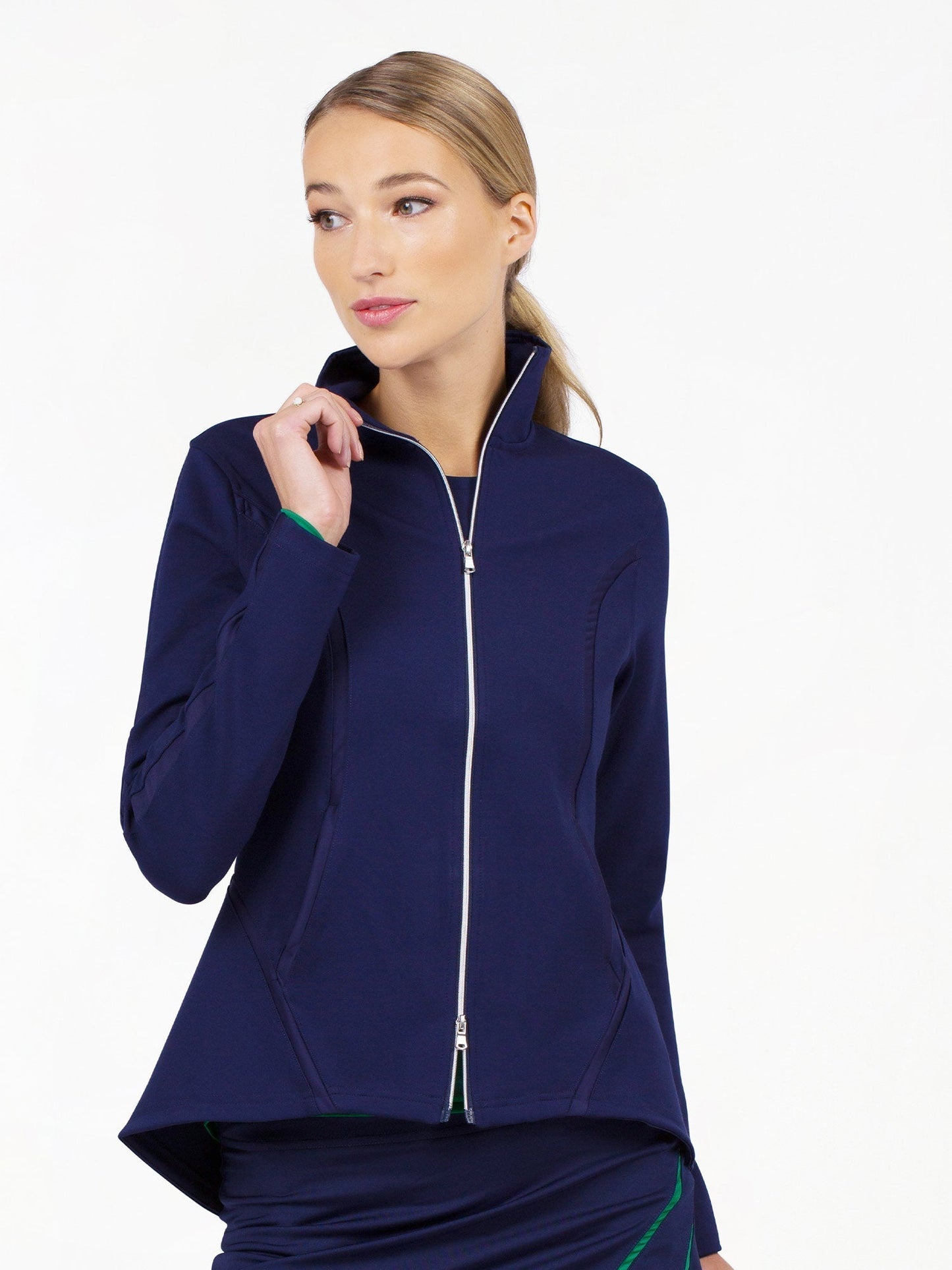 A woman with blonde hair in a low ponytail models the Harlow Knit Jacket - Ink by inPhorm NYC, a navy sun protection jacket with a high collar. She smiles subtly and touches the collar while looking left against a white background.