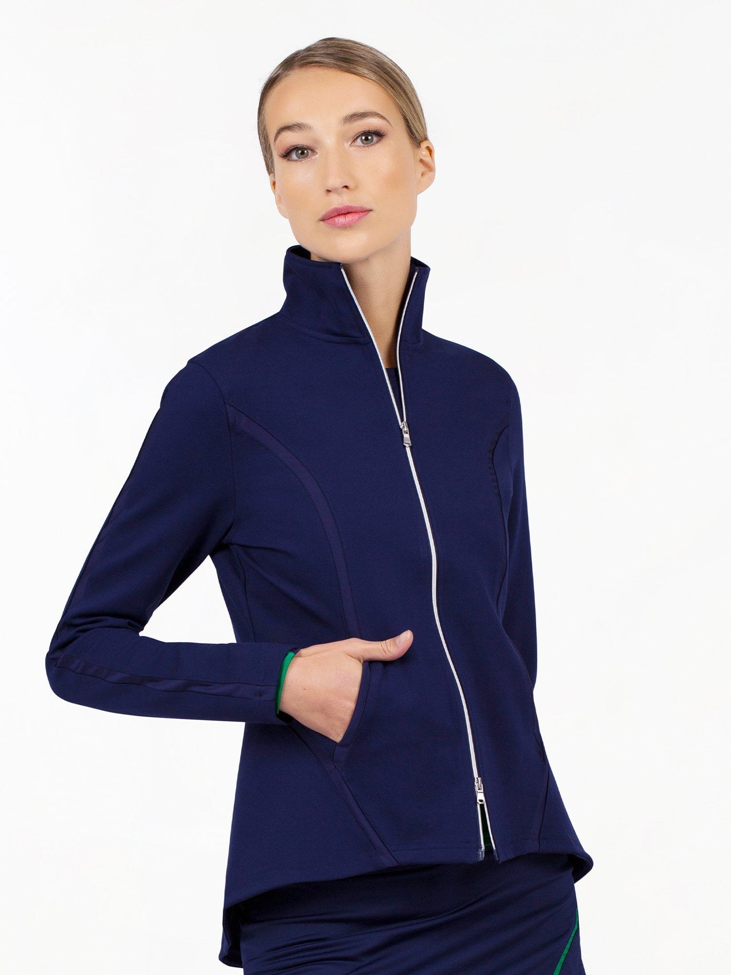 A woman with blonde hair tied back wears the inPhorm NYC Harlow Knit Jacket in Ink, featuring a high collar, silver zipper, and UPF 50+ protection. She stands against a white background, looking to the side with one hand in her pocket.