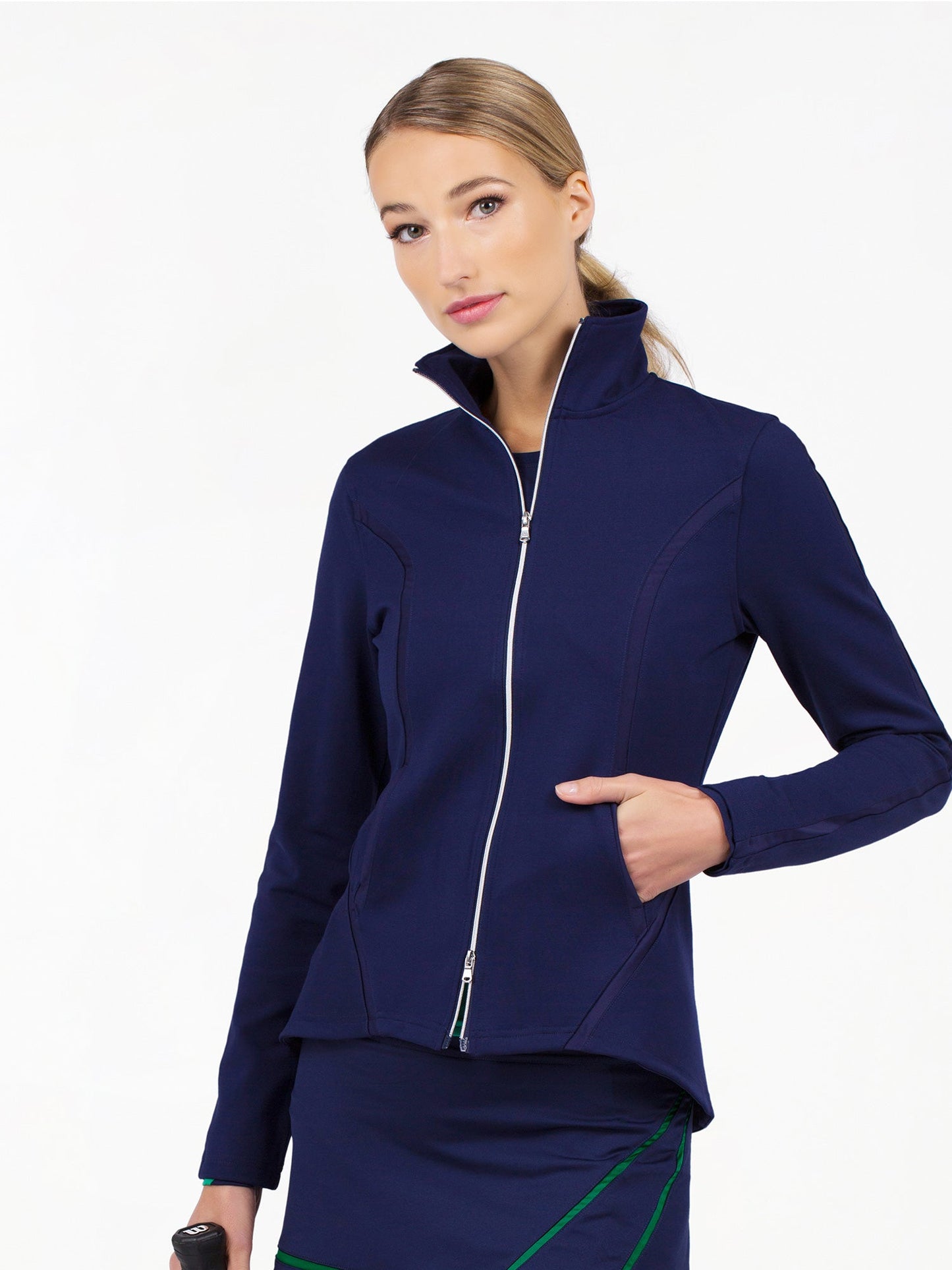 A woman with blonde hair tied back wears the inPhorm NYC Harlow Knit Jacket - Ink, a navy moisture-wicking zip-up with a high collar, and a matching skirt with green accents. She poses against a white background holding a black object.
