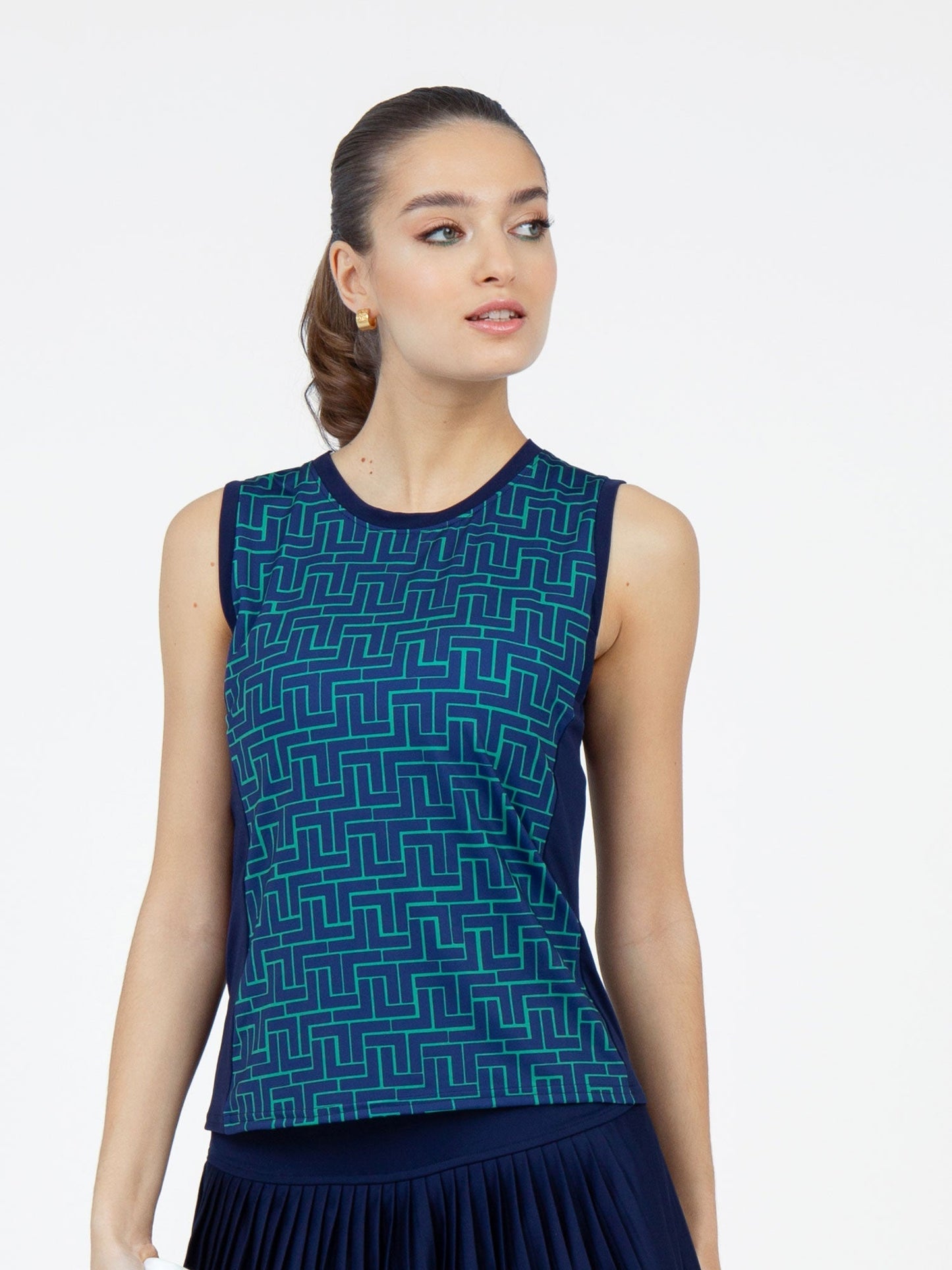 A woman models the inPhorm NYC Harper Performance Tank in Ink Tessel/Ink, featuring a sleeveless, moisture-wicking design and geometric turquoise pattern. She stands against a white background with her hair tied back, looking to her right.