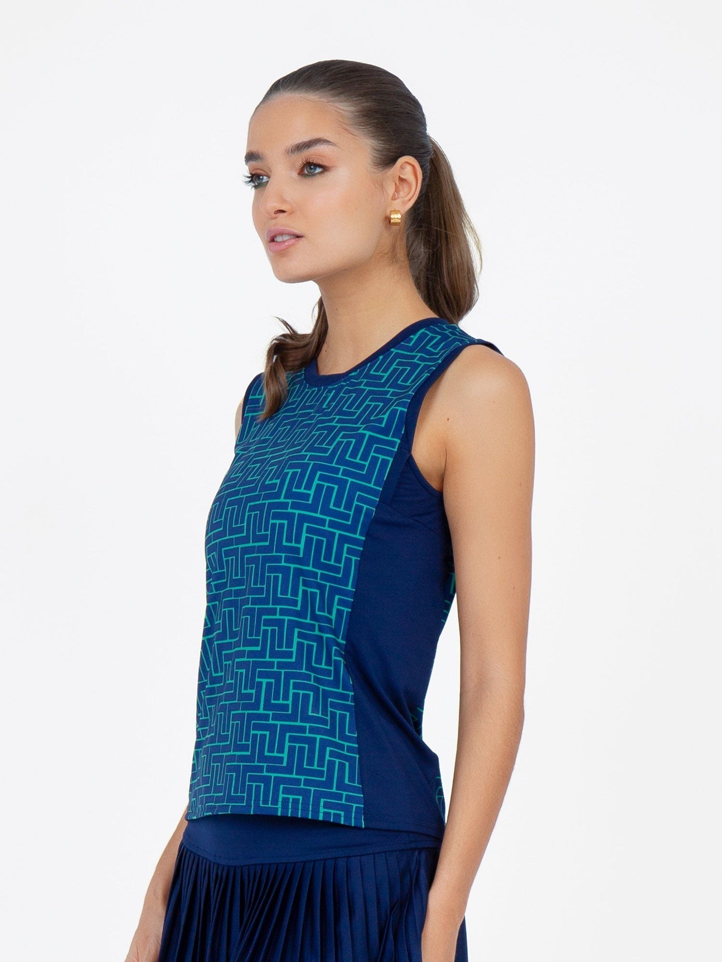 A woman with light skin and brown hair in a low ponytail wears the inPhorm NYC Harper Performance Tank in Ink Tessel/Ink, a sleeveless, navy blue relaxed-fit top with a geometric teal pattern. She stands against a white background, looking slightly left.